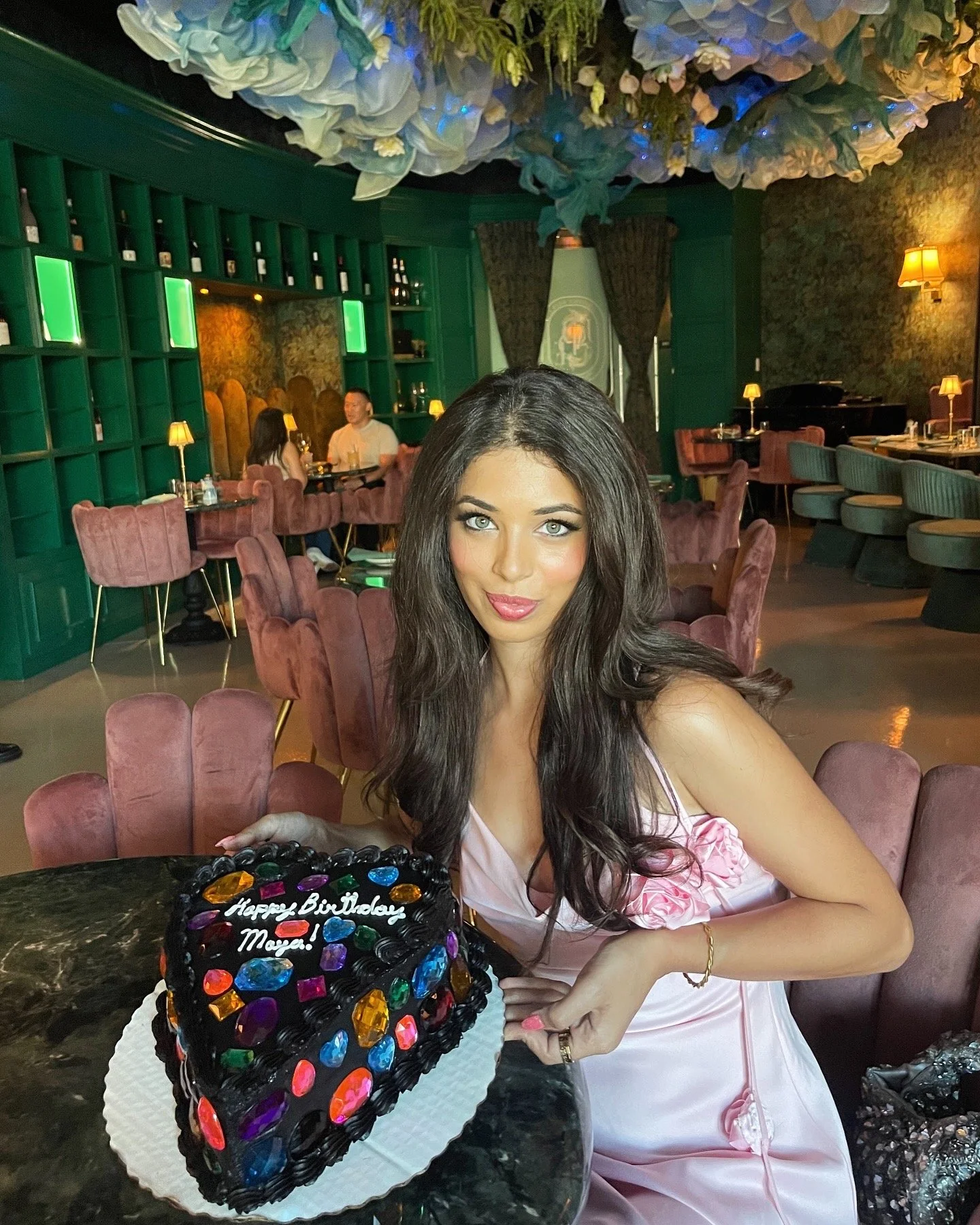 A woman with long dark hair and light skin in a pink dress sitting at a table in a decorated restaurant with a colorful cake that has 'Happy Birthday Maya!' written on it.