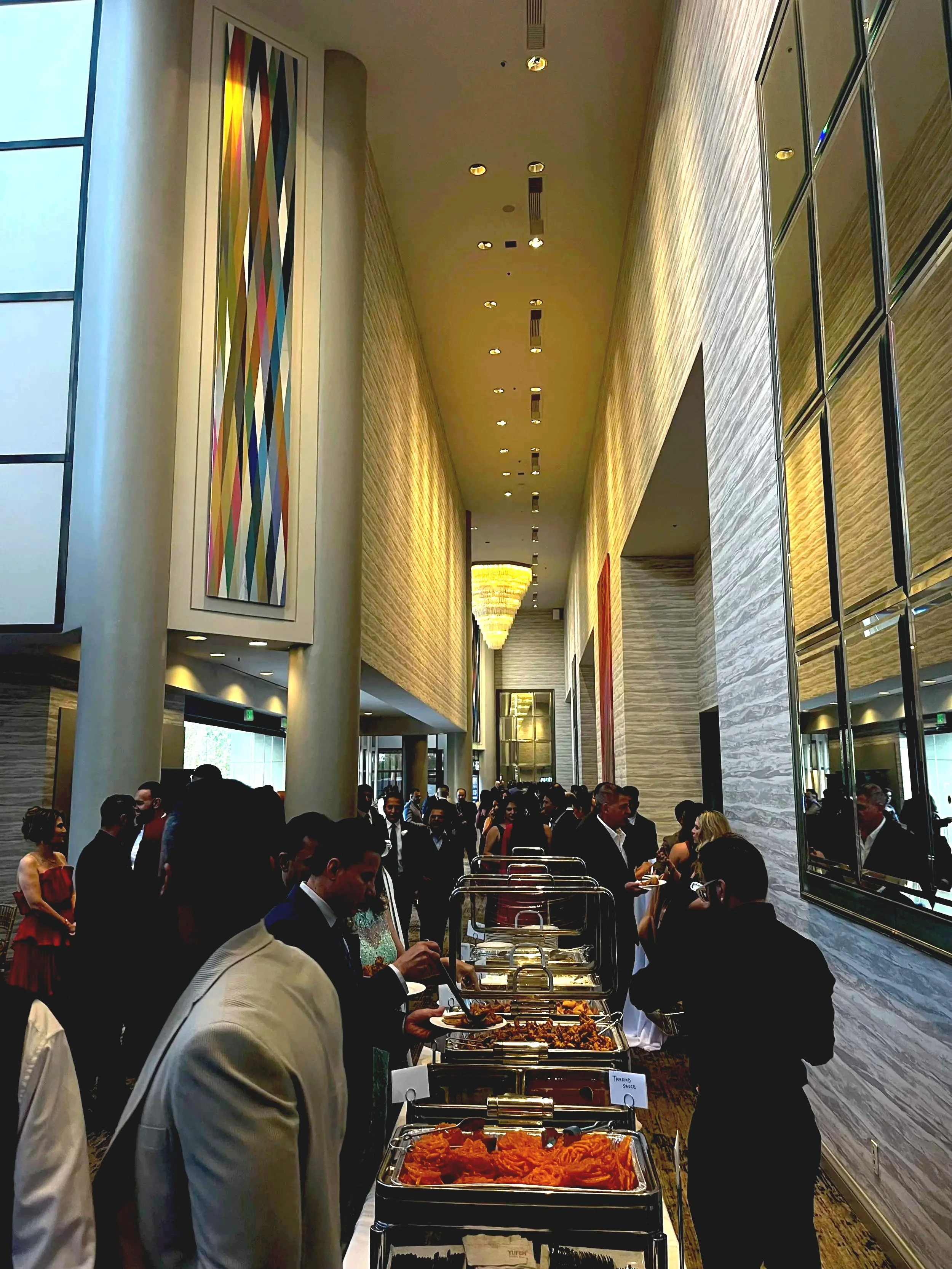 People at a buffet table in a high-ceiling modern hotel lobby. More people stand in line behind the table.