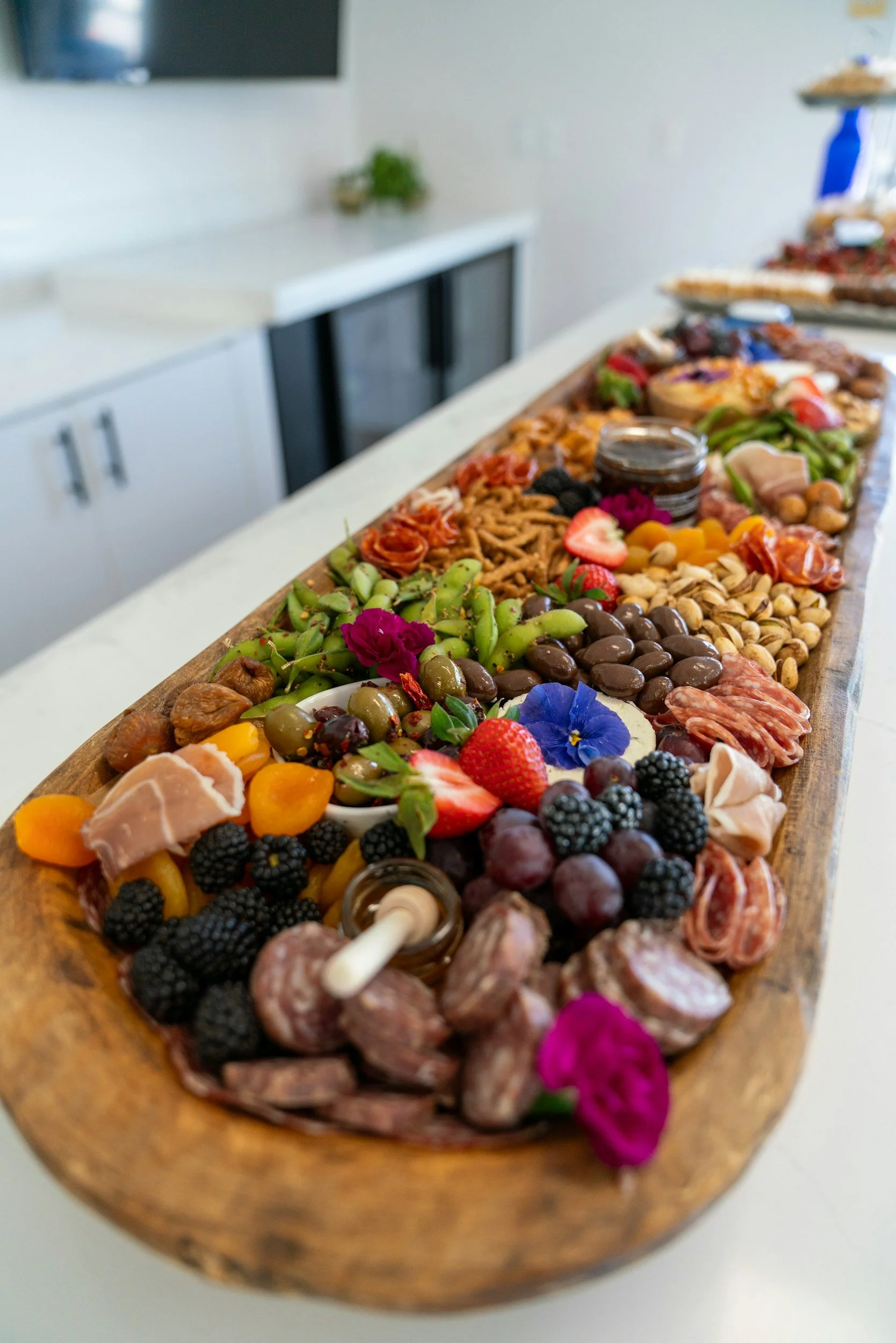 A long wooden board filled with a variety of colorful fruits, nuts, chocolates, cheese, and cured meats, decorated with edible flowers.