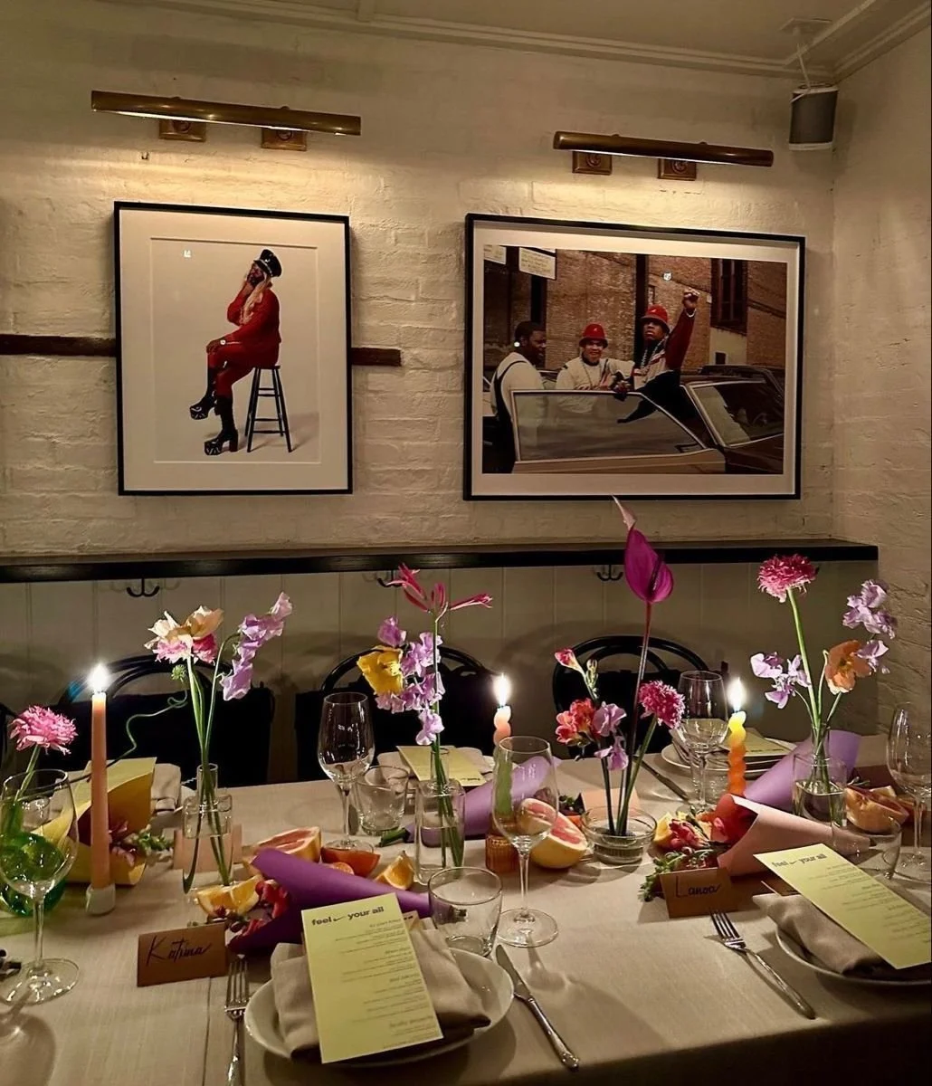 A decorated dining table with flowers, candles, and place cards, set for a celebration, with framed photographs on a white brick wall in the background company dinner