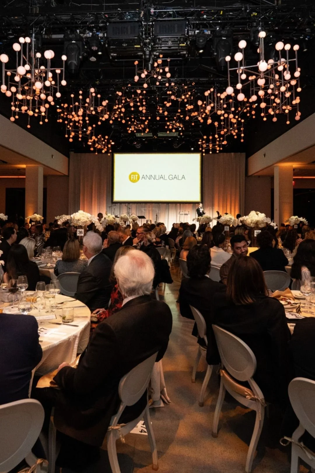 An audience seated at round tables, attending an annual gala event with a large stage, floral centerpieces, and a screen displaying 'FIT Annual Gala'.