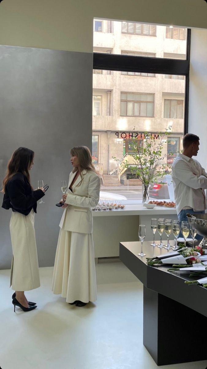 Two women are attending an indoor social event, having a conversation while holding champagne glasses. One woman has dark hair and is wearing a dark jacket with cream wide-leg pants, and the other has blonde hair and is dressed in a cream blazer and matching long skirt. There is a man in the background on the right, facing away, with a white shirt. A window behind them shows an urban street scene, and a table with snacks and drinks is visible along with a vase with flowers.