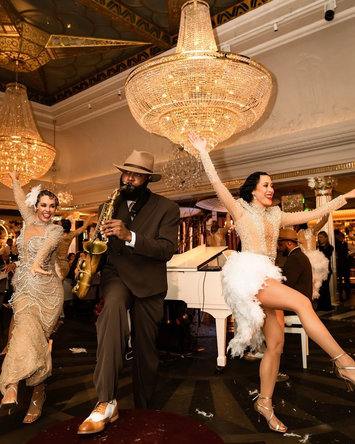 Performers dancing and playing saxophone at an elegant event with chandeliers and a grand piano in a lavish setting.