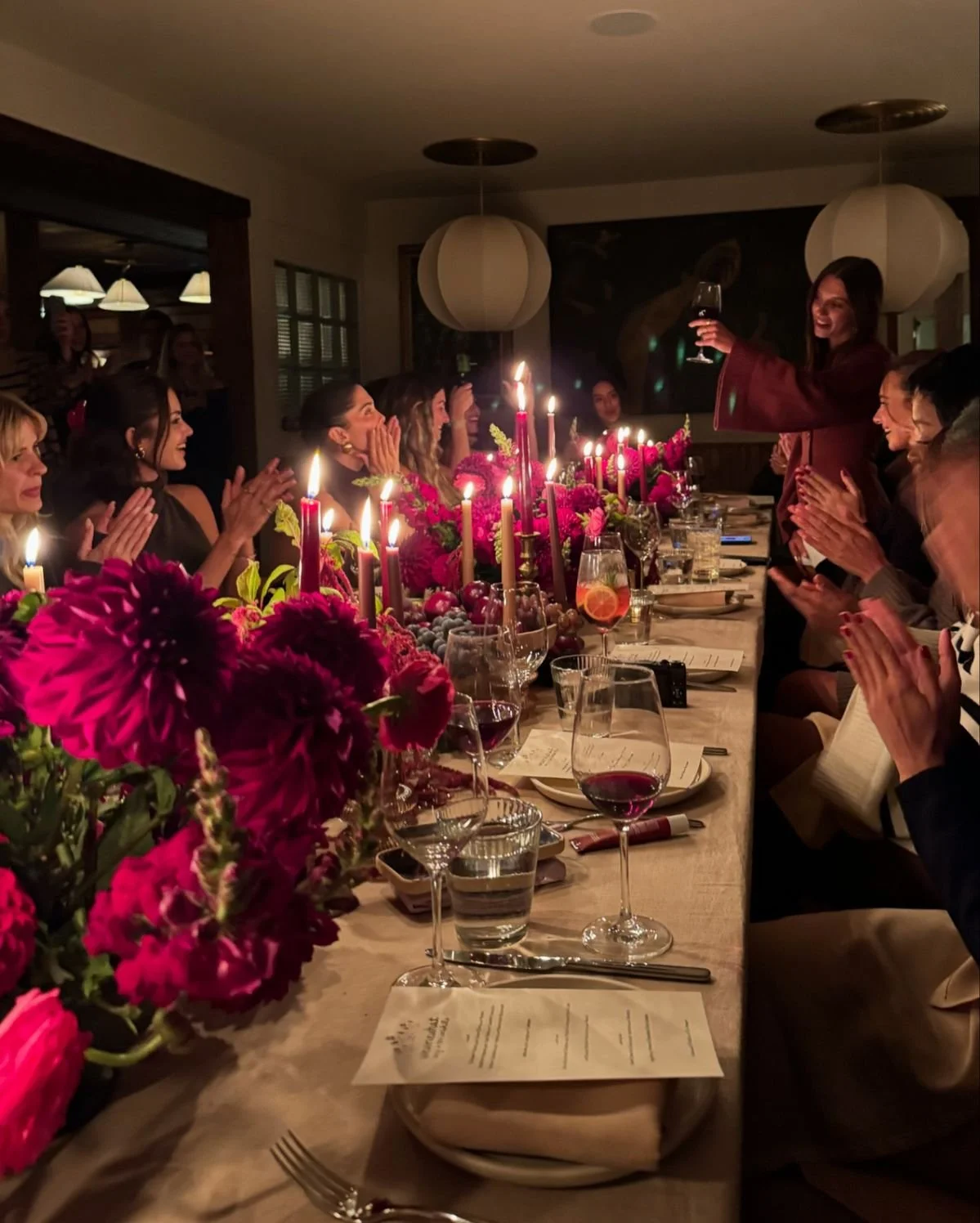People gathered around a long dinner table decorated with pink flowers and candles, celebrating a birthday. One woman is standing and toasting with a glass of wine, while others are clapping and smiling.