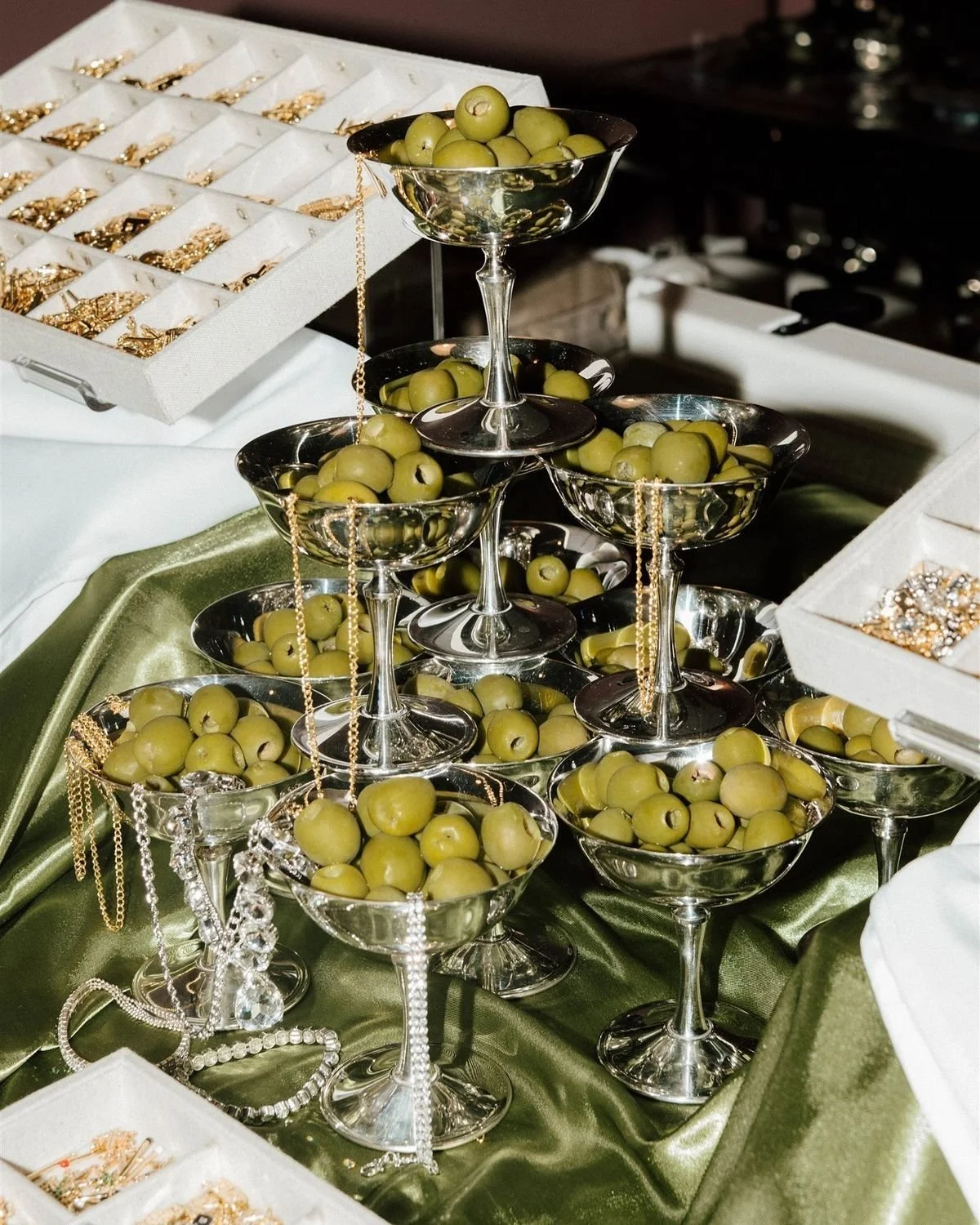 Multiple silver bowls filled with green olives displayed on a table with a green satin tablecloth, with jewelry and jewelry boxes nearby.