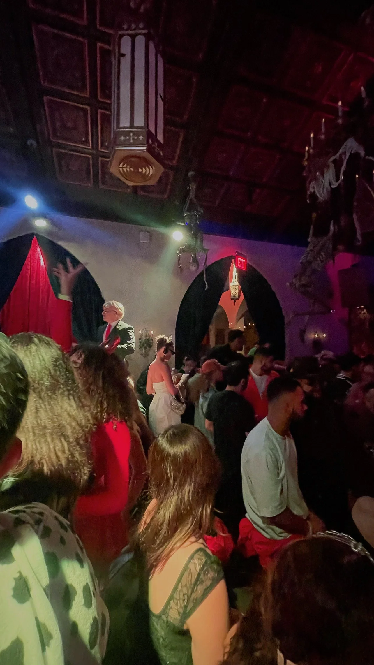 A crowded indoor event with people standing and sitting, a person on stage, and Halloween decorations hanging from the ceiling.