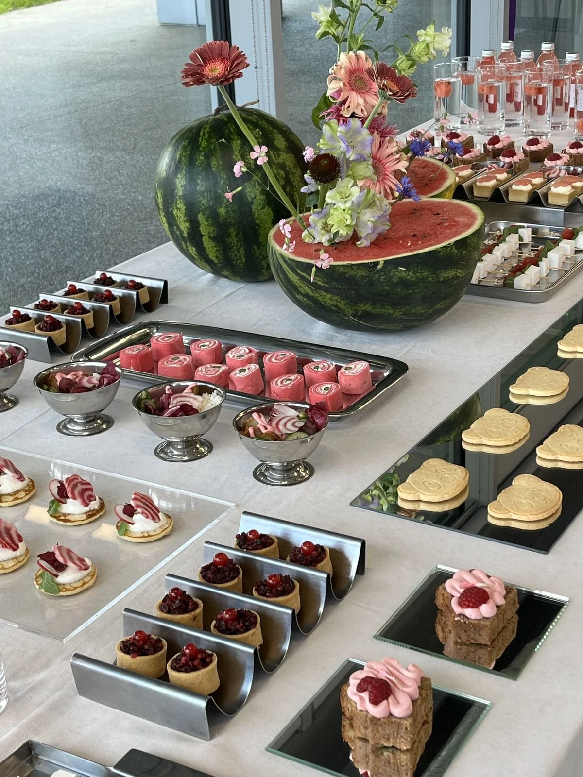 Display of assorted desserts and watermelon arrangements at a dessert table, with fresh flowers and glasses of water in the background.