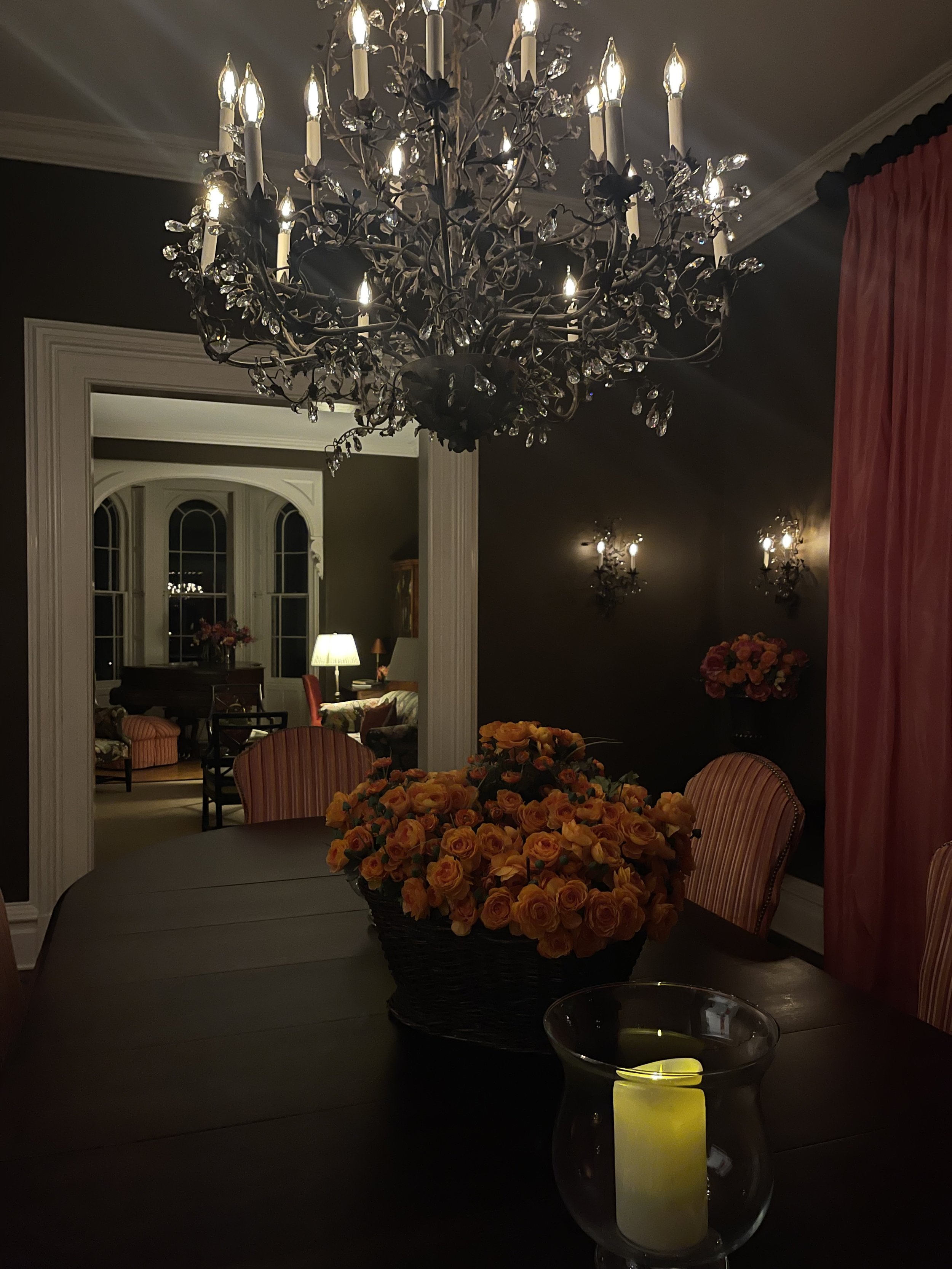 A dining room with a large chandelier, a table with a flower centerpiece, a candle in a glass holder, and a dark wall with wall sconces.