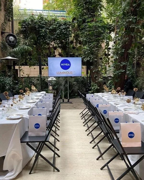 Outdoor event space with long tables covered in white tablecloths, set for a meal with plates, glasses, and floral centerpieces. Black chairs are arranged on both sides of the tables, and a large screen displays the NIVEA logo along with the word LUMINOUS in the background, surrounded by greenery and trees.