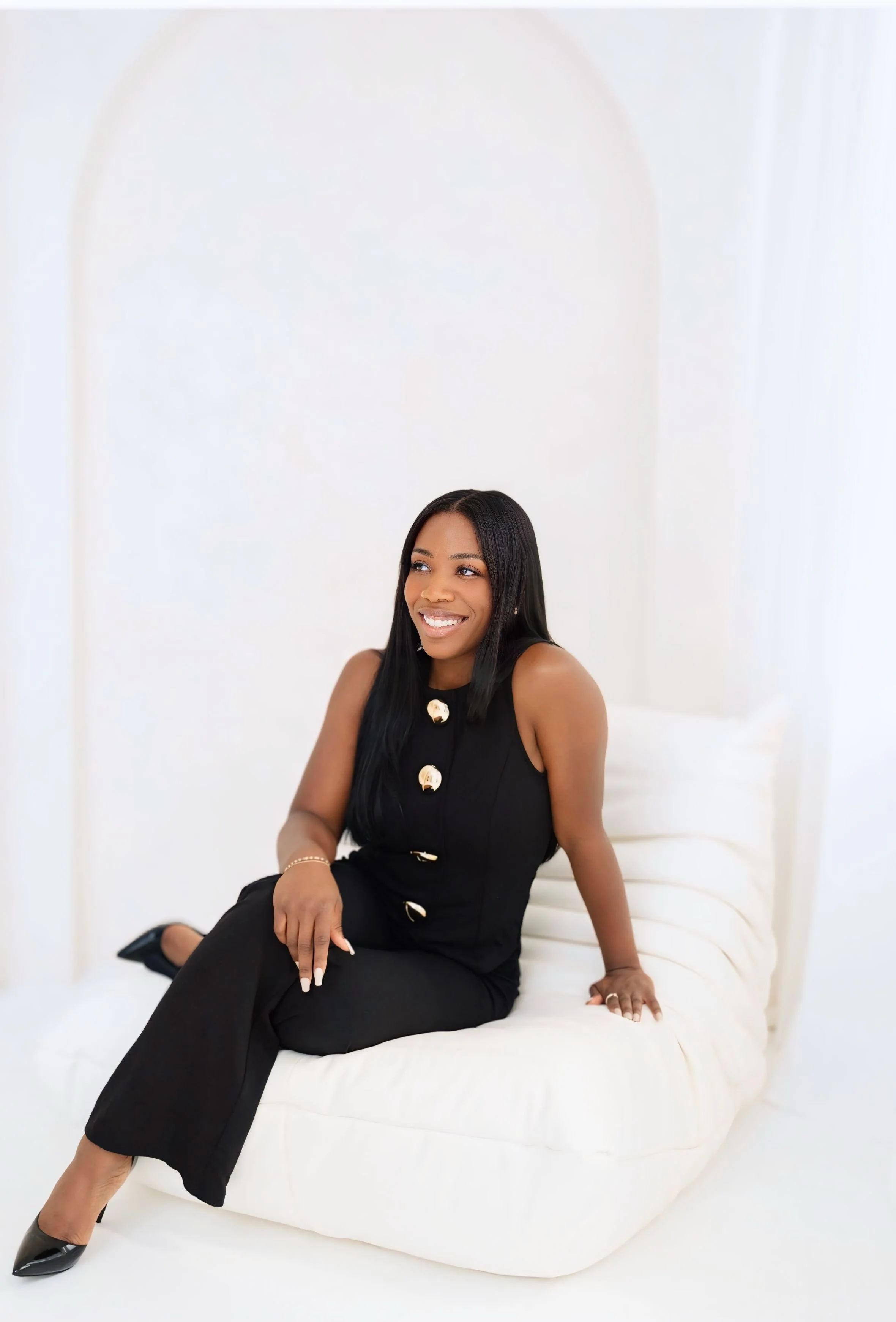 Founder of Olive Wellness Concierge Services seated in a minimalistic studio wearing black sleeveless outfit with gold buttons.