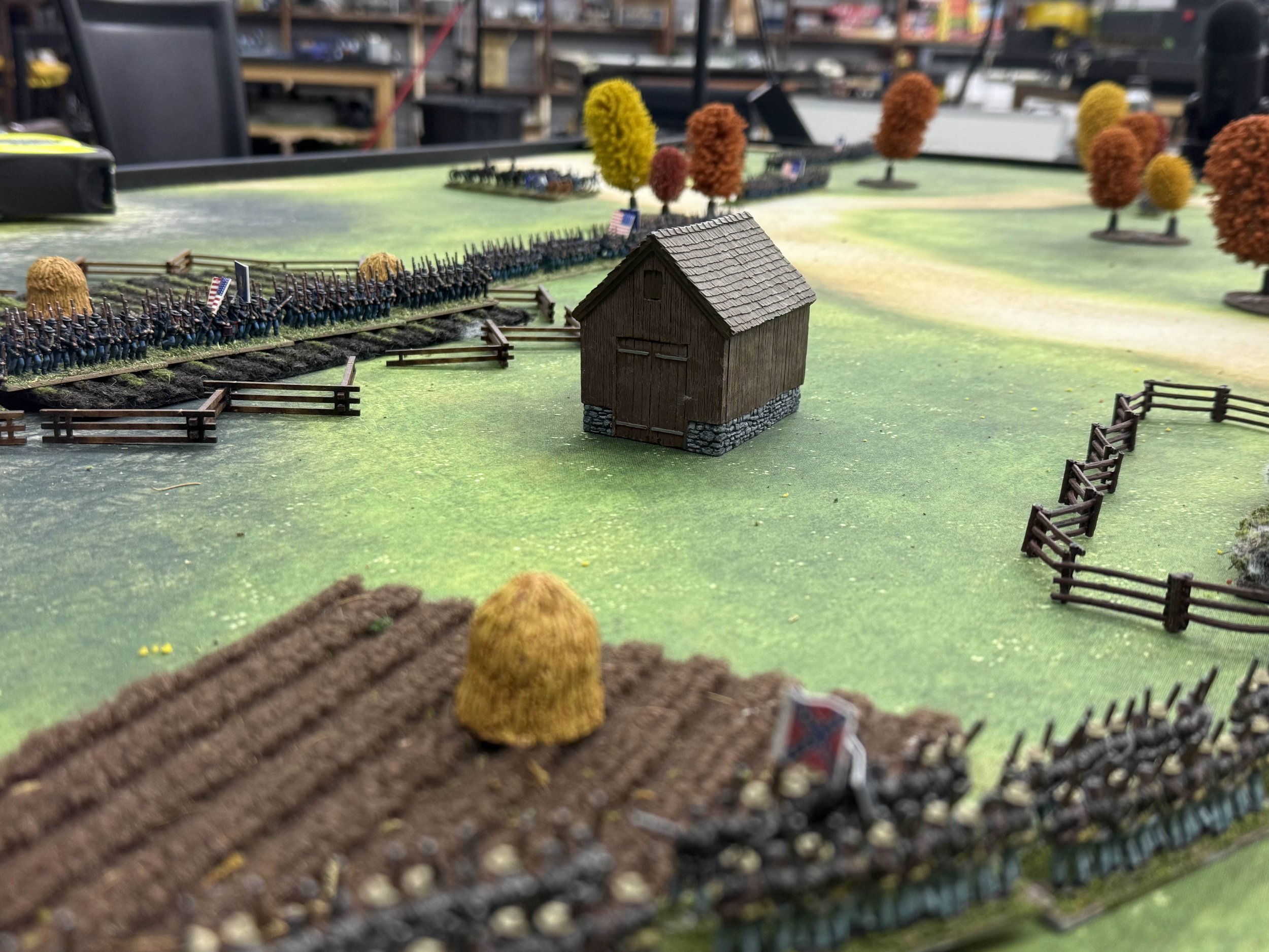 Miniature battlefield with toy soldiers arranged in formation, a small barn, and miniature trees scattered across a green landscape.