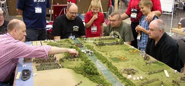 People gathered around a tabletop war game with miniature terrain and figures.