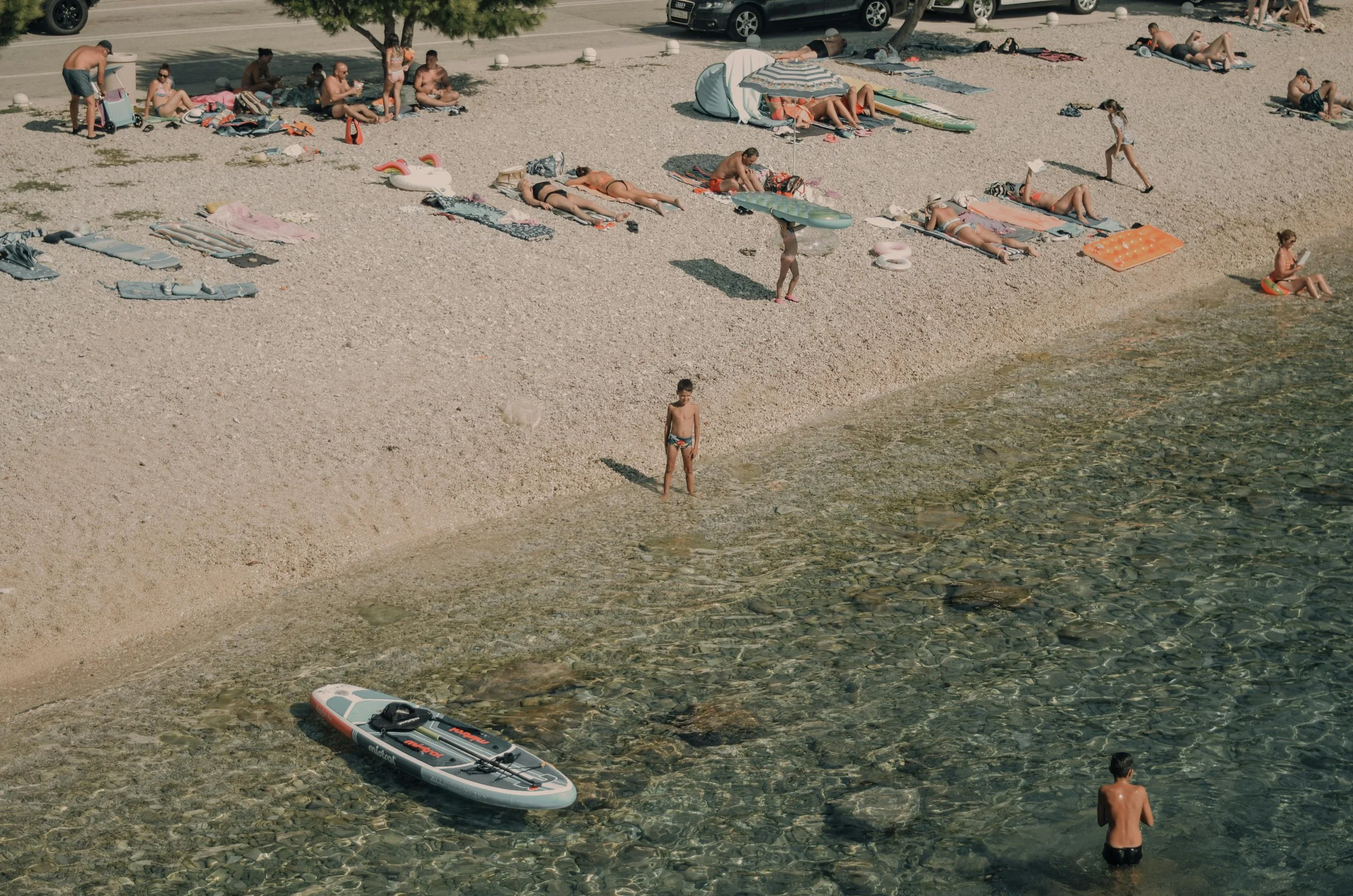 Beach scene with people sunbathing, sitting, walking, and swimming along the shoreline, with an inflatable paddleboard in the shallow water and cars parked beyond the sandy beach.