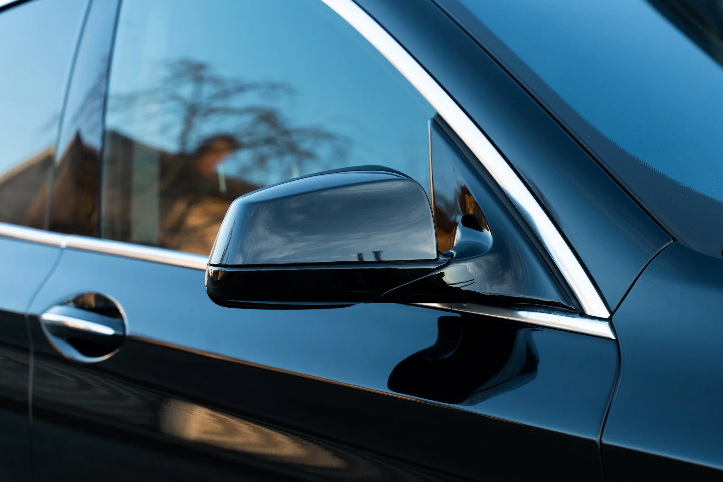 Close-up of a black car door with a side mirror and window, reflecting the sky and trees.