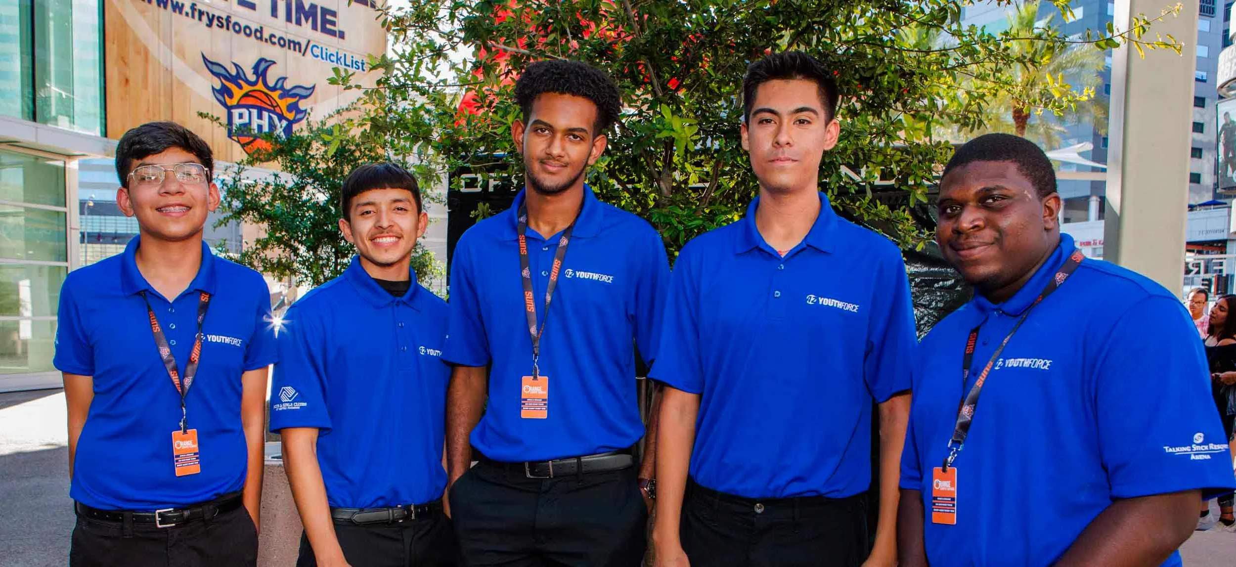 Group of five young men wearing blue shirts with 'YOUTHFORCE' logos, standing outdoors in front of greenery with a city background, smiling at the camera.