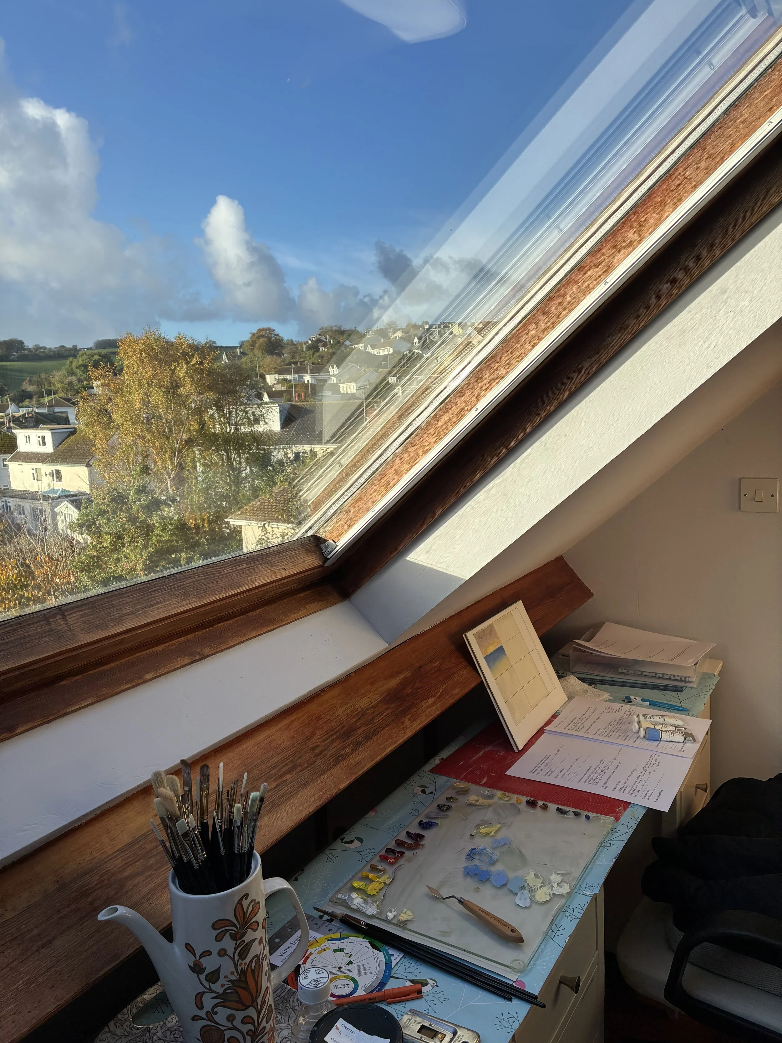 Fi Mackenzie's Studio in Brixham with art supplies laid out, including a container with paintbrushes, a palette with mixed paint, and papers with drawings. The window shows views over Brixham's green fields.