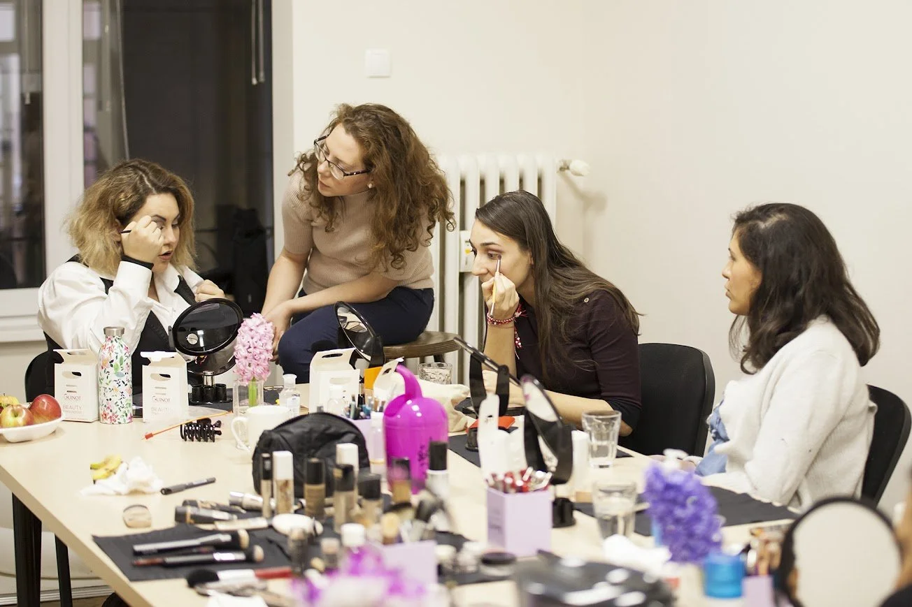 Four women are gathered around a table with makeup products, mirrors, and beauty supplies, one is applying makeup while others are watching or talking.