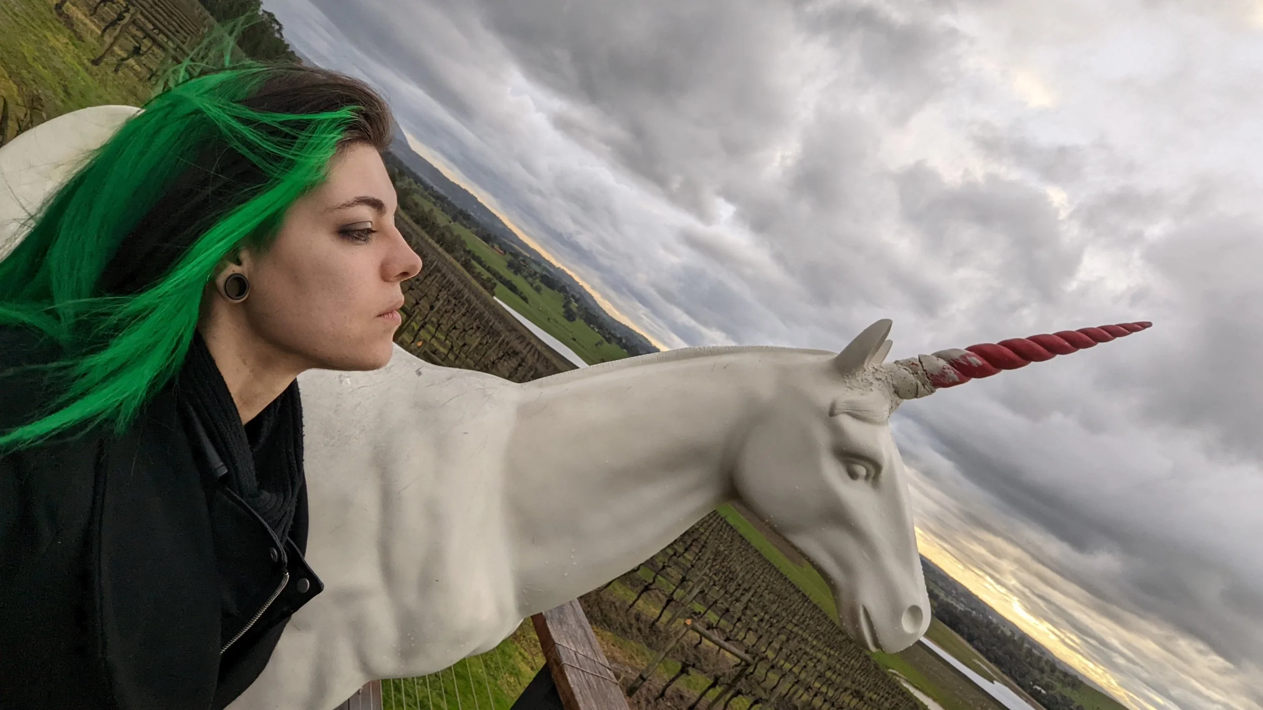A woman with green and black hair and large gauge earrings taking a selfie next to a white unicorn statue with a red and white spiral horn, against a cloudy sky and vineyard landscape.