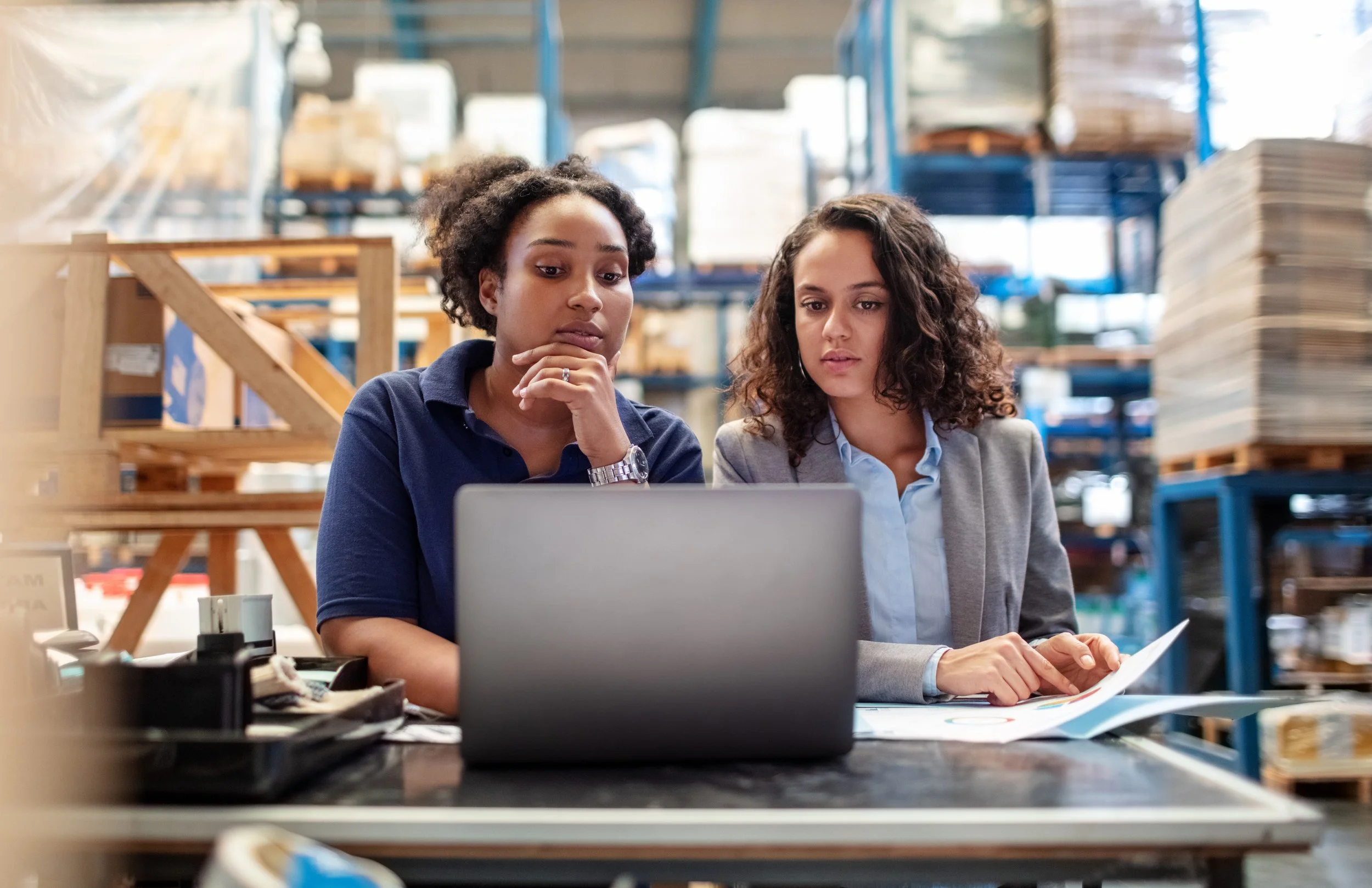 Two women working together at a desk in a warehouse, looking at a laptop and documents.