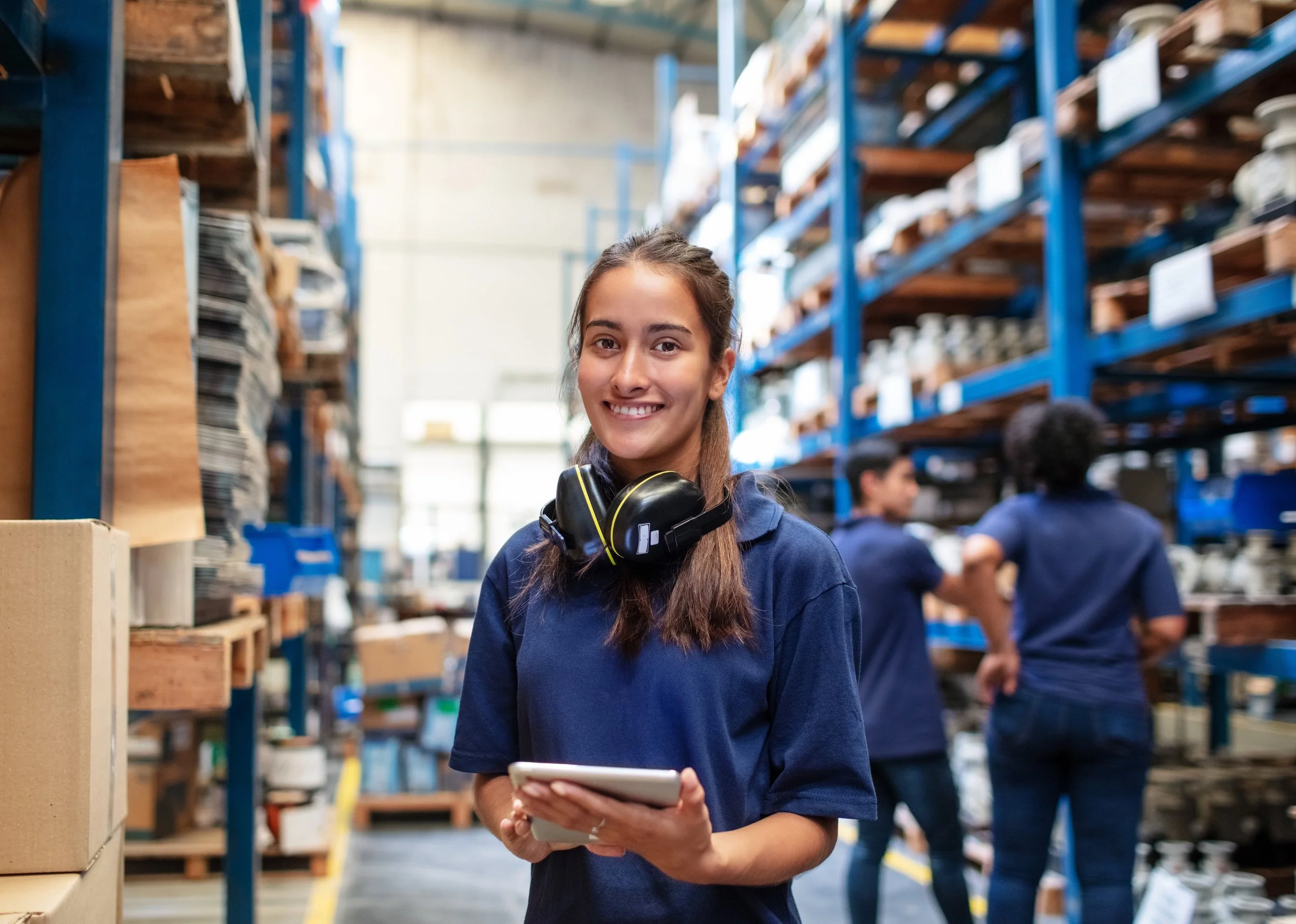 A young woman smiling while holding a tablet in a warehouse or industrial storage area, with shelves of materials and other workers in the background.