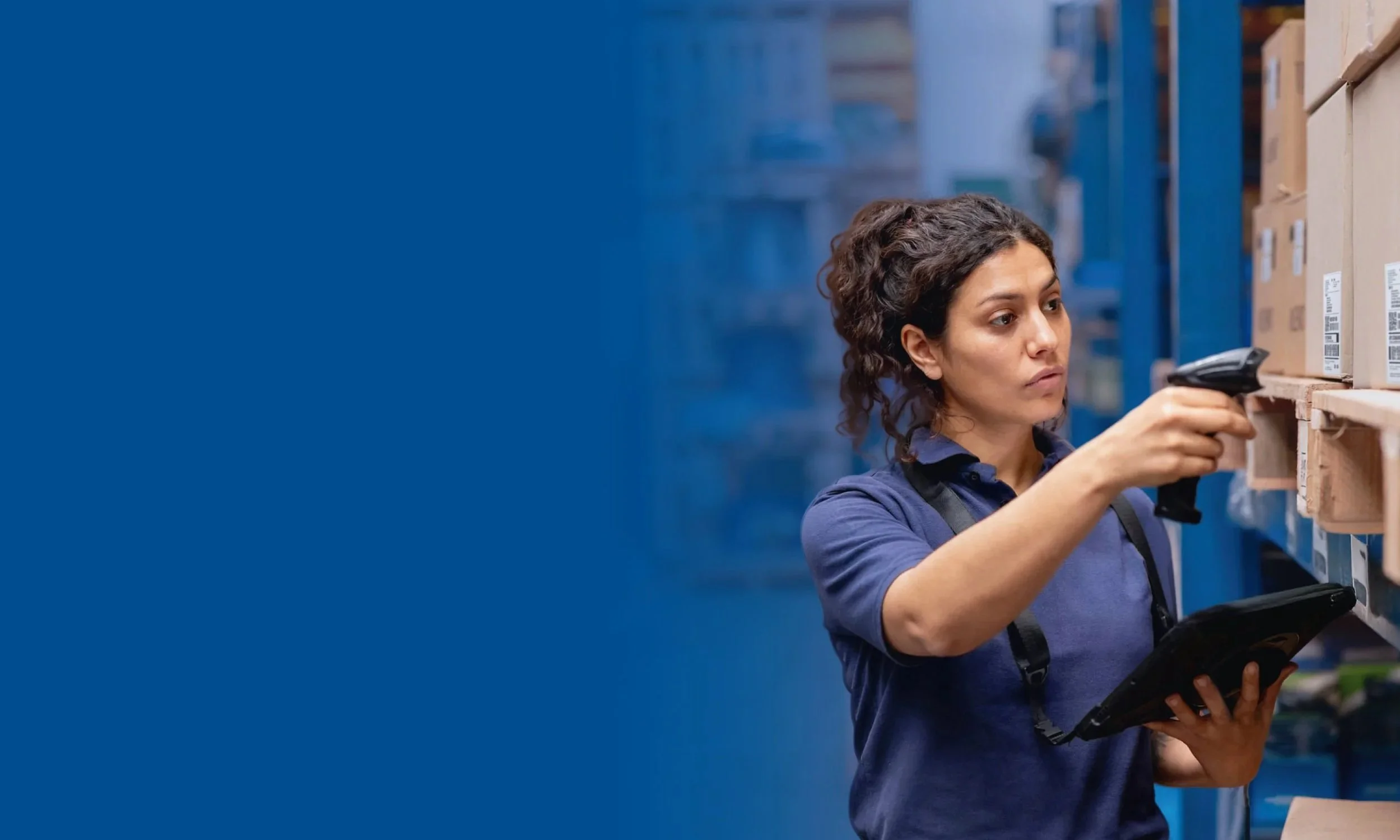 A woman with curly hair wearing a blue shirt and a black apron in a warehouse, scanning a barcode on a cardboard box with a handheld scanner.