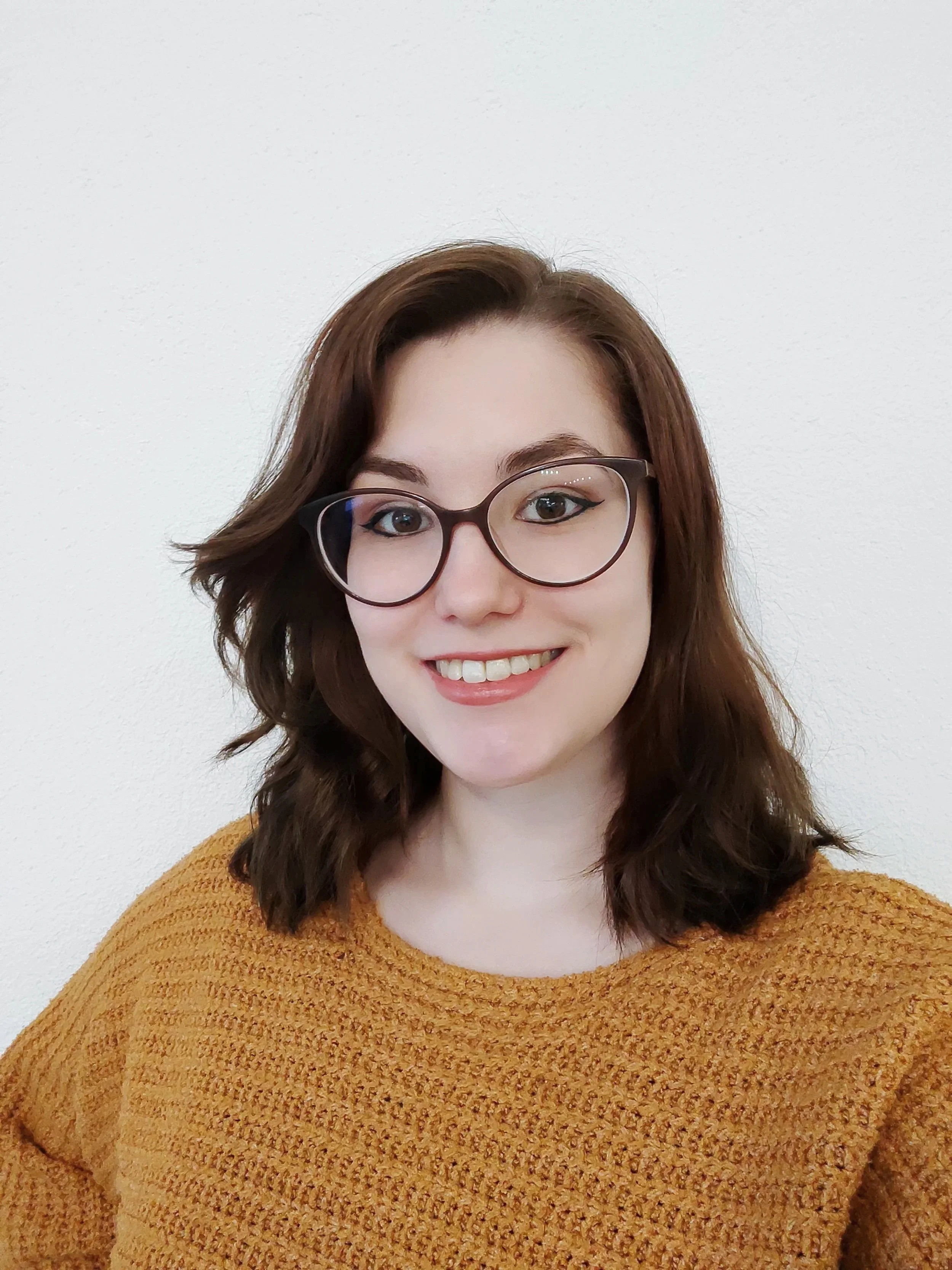 Junge Frau mit braunen Haaren, großen Gläsern und braun-orangefarbenem Pullover vor weißer Wand.