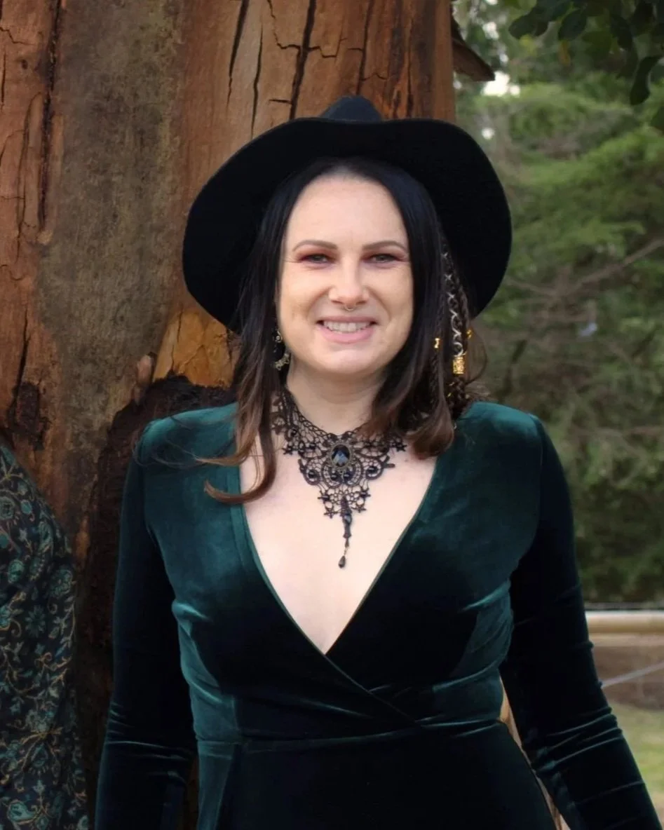 A woman with dark hair, wearing a black wide-brimmed hat, ornate black necklace, and a deep green velvet dress, standing outdoors in front of a tree and green foliage.