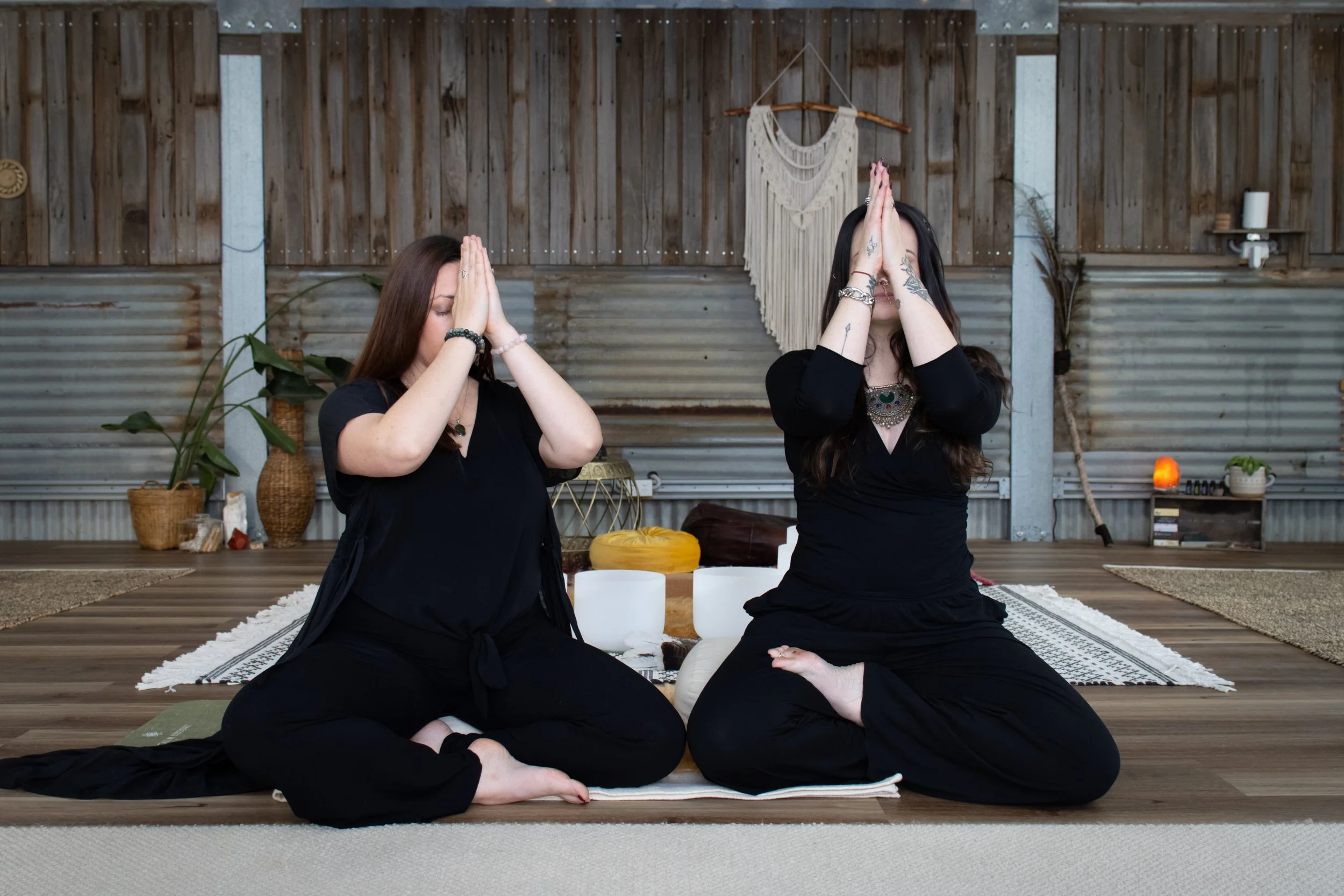 Two women practicing yoga in a rustic wooden room, sitting cross-legged on a rug, with their hands pressed together in a prayer position in front of their faces.