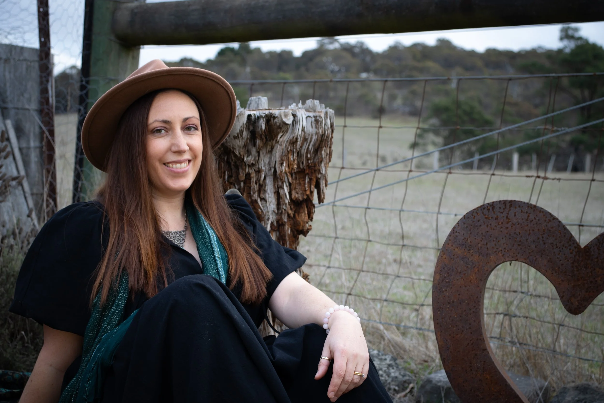 A woman sitting outdoors near a fence with a rusty heart-shaped sculpture, wearing a brown hat and black dress, smiling.