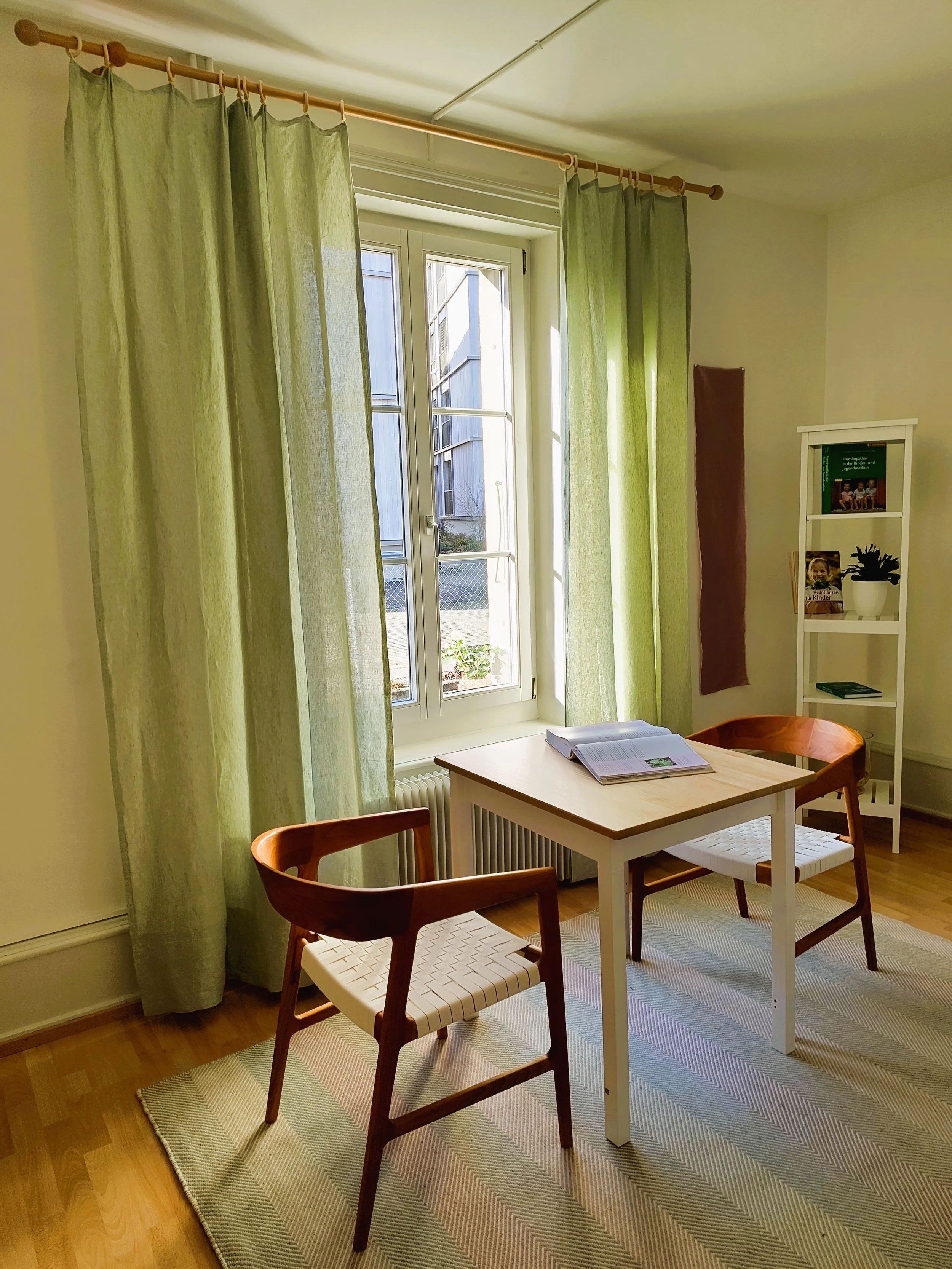 Ein heller Raum mit einem großen Fenster, grünen Vorhängen, einem weißen Tisch, zwei Holzhockern, einem roten Stuhl, einem Bücherregal und einem Teppich.