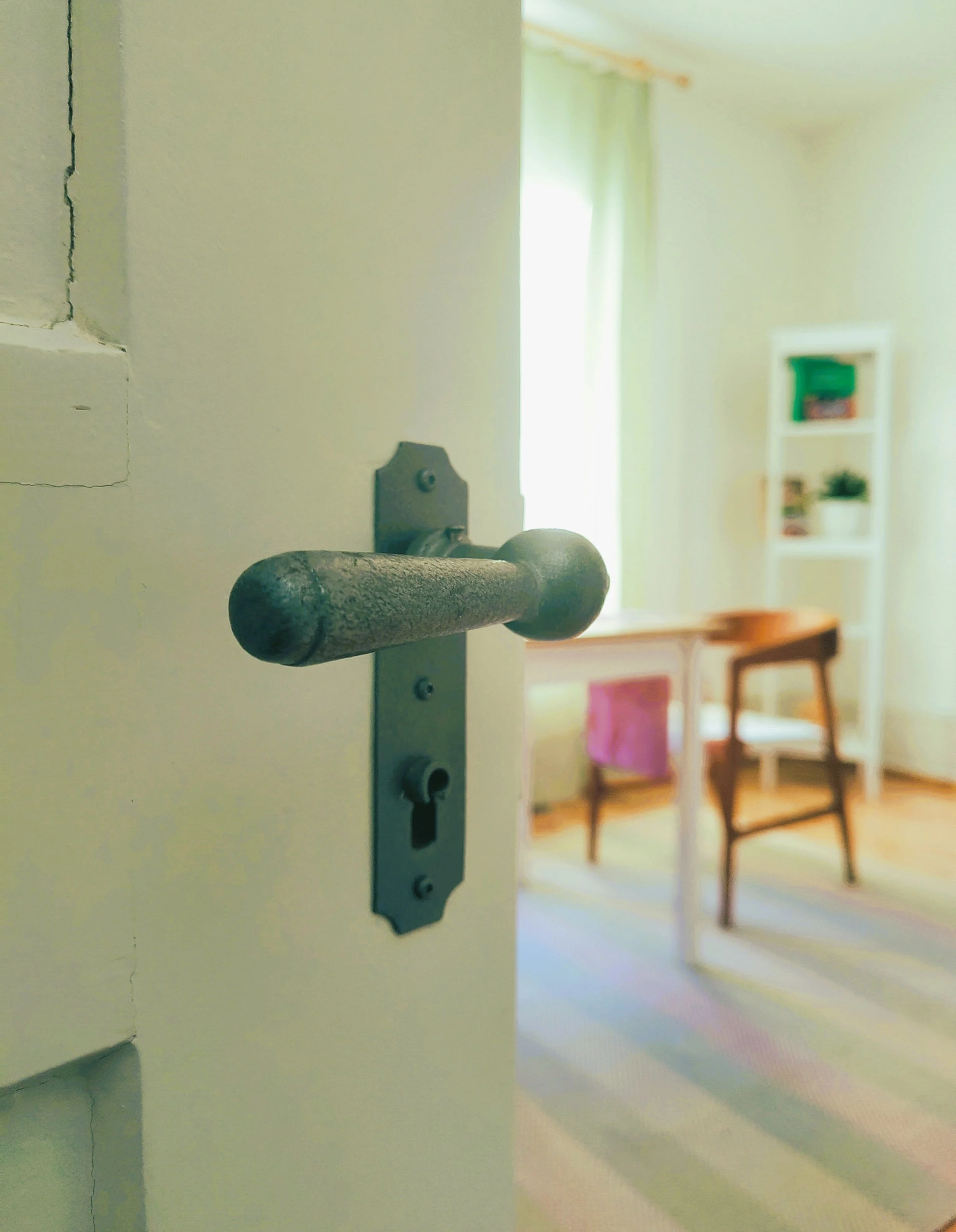 Eine Tür mit einem abgenutzten Eisenriegelverschluss, geöffnet in einen hell erleuchteten Raum mit Möbeln und Vorhängen.