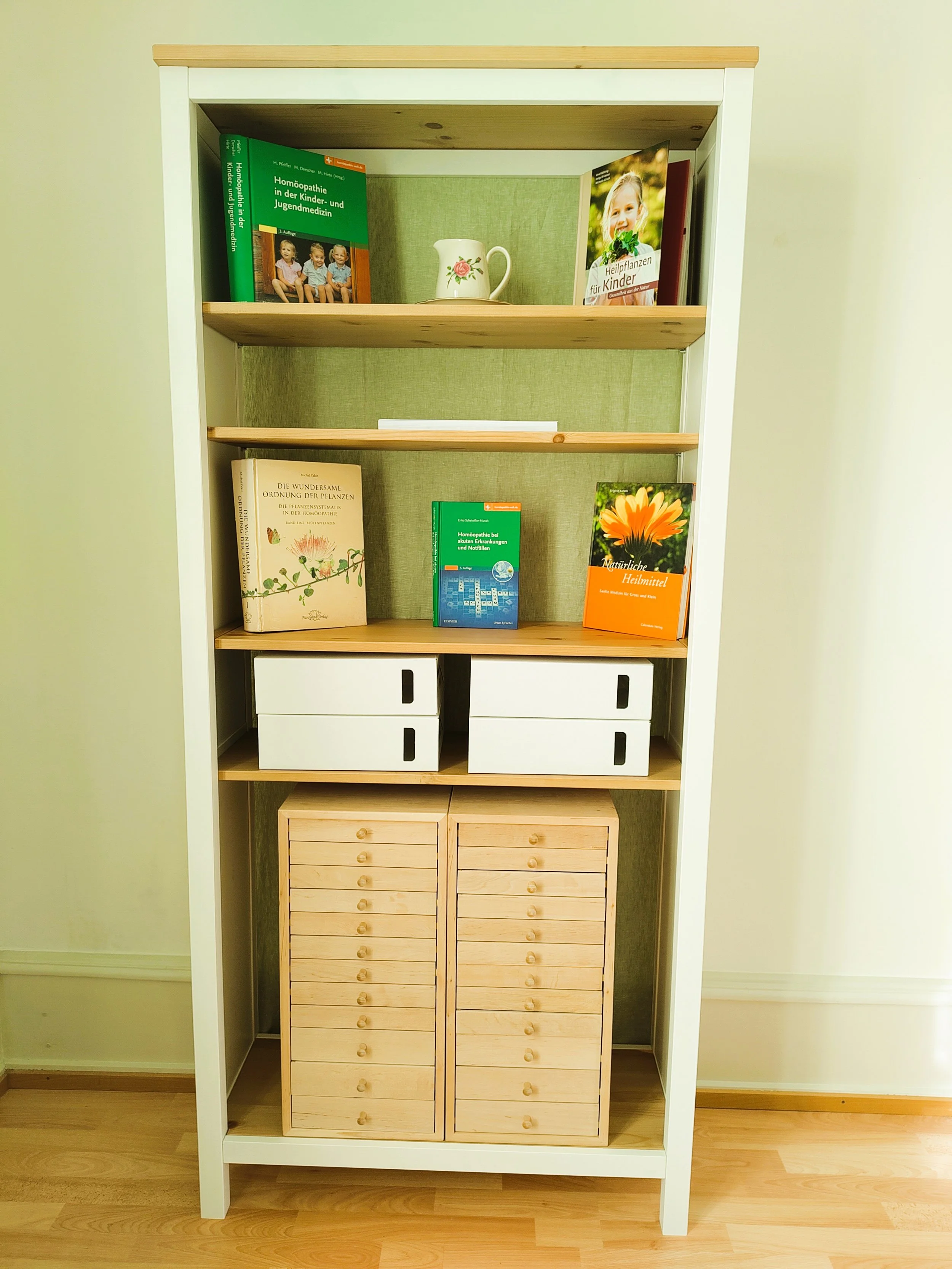 Ein Bücherregal mit mehreren Fächern, darin liegen Bücher, weisse und schwarze Boxen, sowie eine Holzschachtel mit mehreren kleinen Schubladen. Auf den oberen Fächern befinden sich Bücher, eine Vase und eine Tasse.