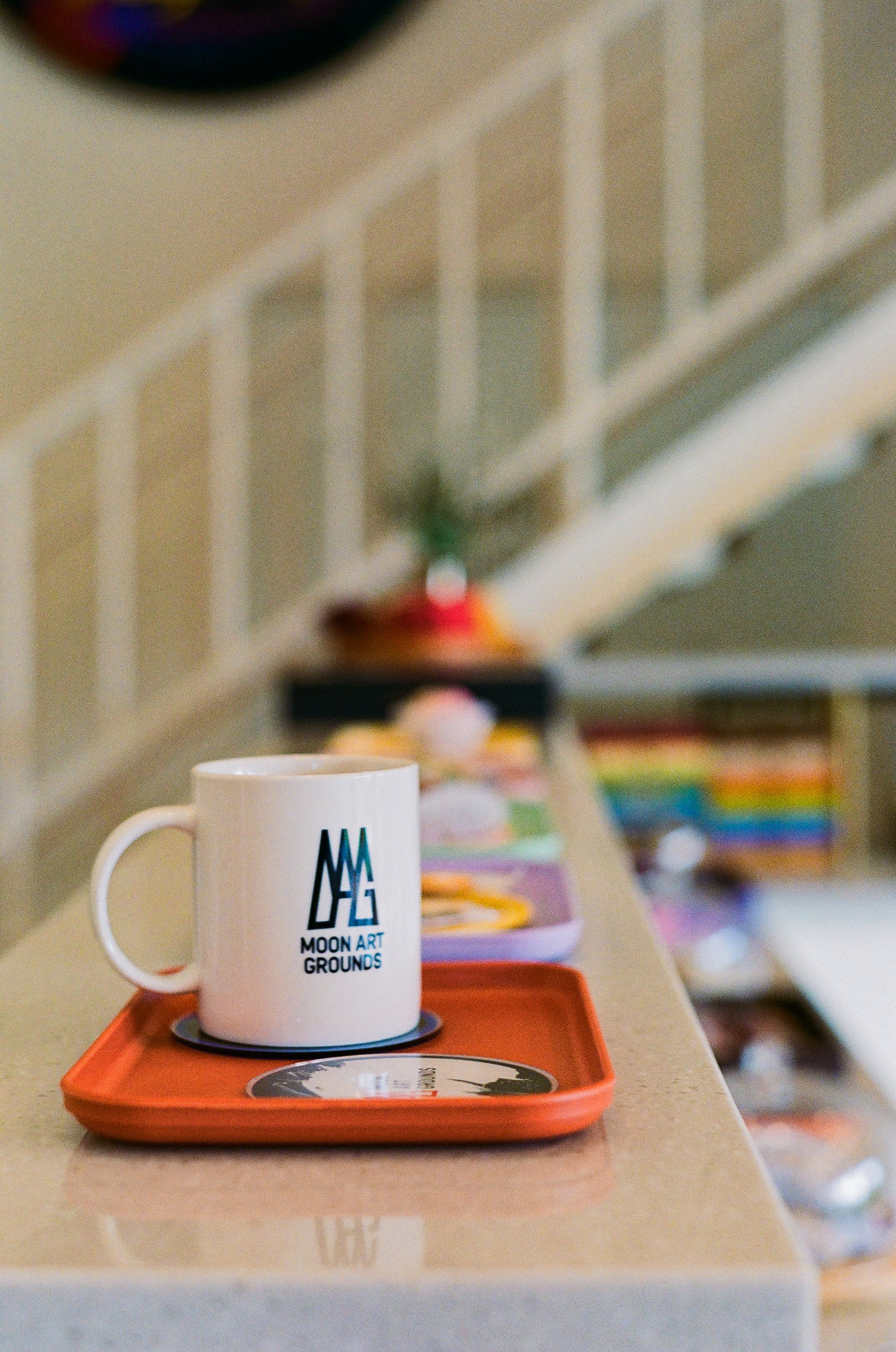 흰색 머그잔이 파란색과 검은색 텍스트로 'MOON ART GROUNDS'라고 쓰여 있으며, 빨간색 접시 위에 놓여 있고, 배경에는 여러 가지 색상의 책과 장식이 보임.