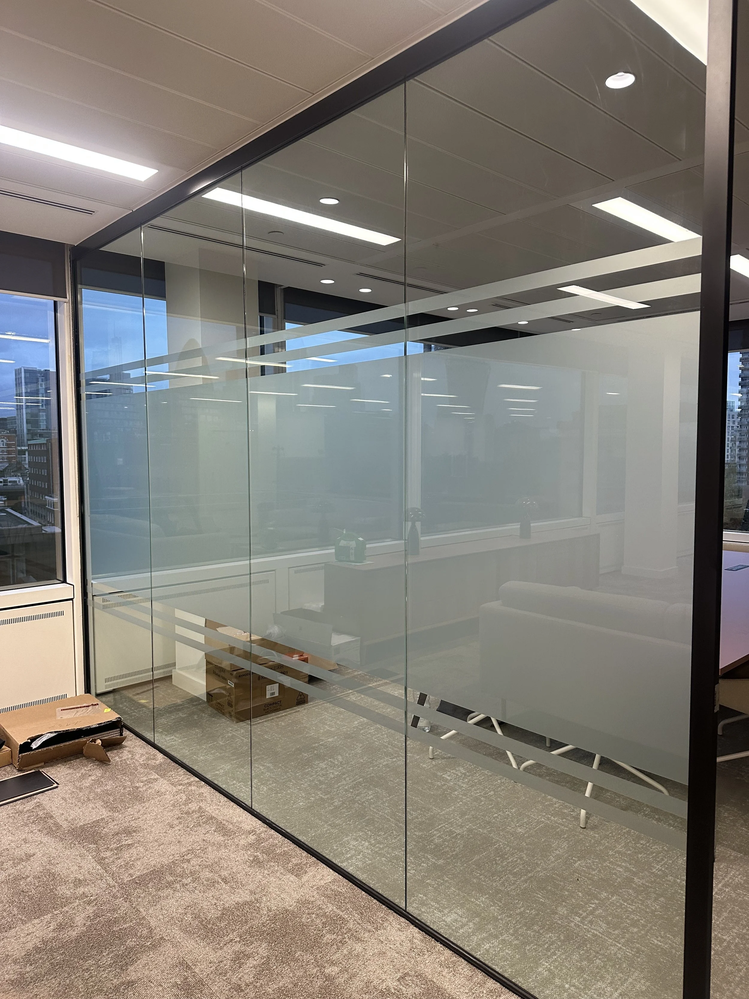 An office space with frostedglass walls and a conference room inside. The room contains a desk, chairs, and some office supplies. The office is well-lit with ceiling lights, and through the windows, a cityscape with tall buildings is visible outside.