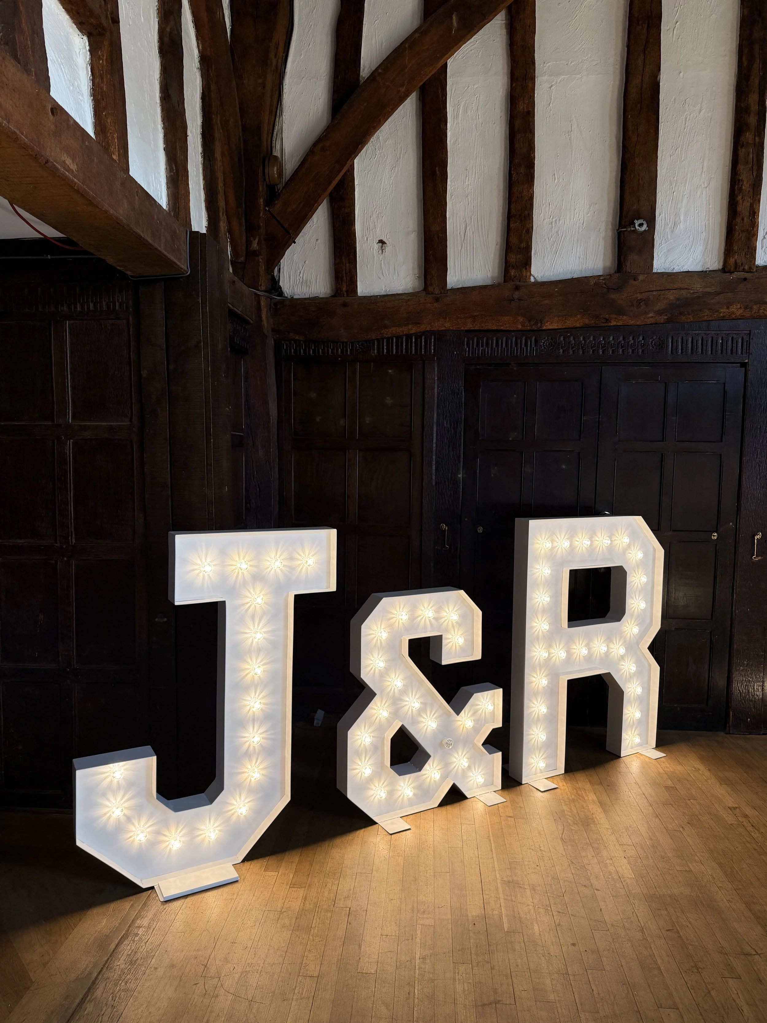 Large illuminated letters spelling out 'J&R' in white with light bulbs inside, positioned on a wooden floor against a dark wood-paneled wall with exposed ceiling beams.