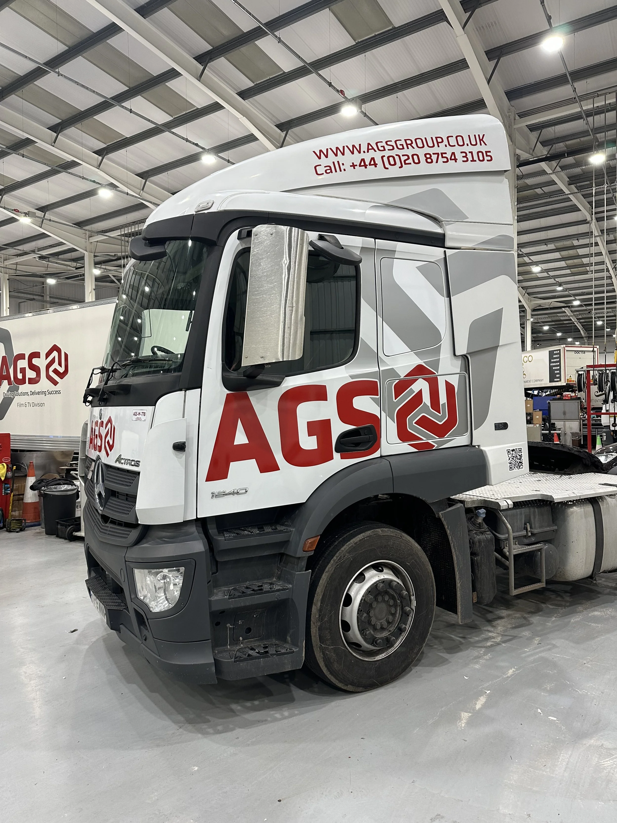 White commercial truck inside a warehouse, with company logo 'AGS' on the side and website and phone number on the upper part of the cab.