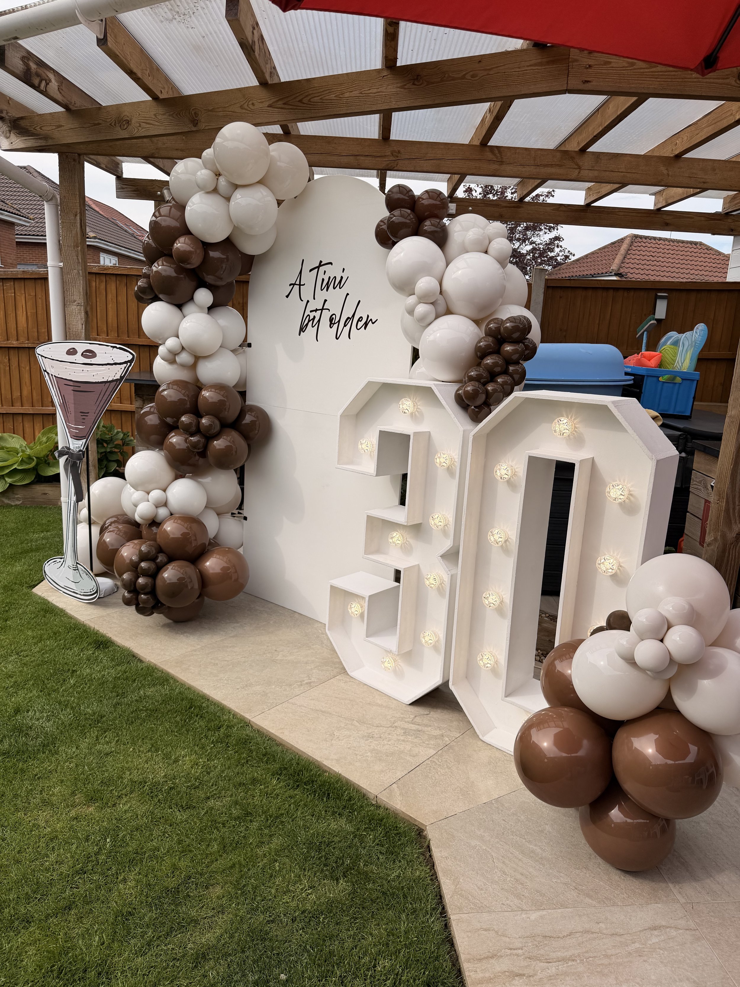 Outdoor birthday celebration decoration with white and brown balloons, a large white board with text, a cutout illustration of a martini glass, and freestanding LED numbers '30'