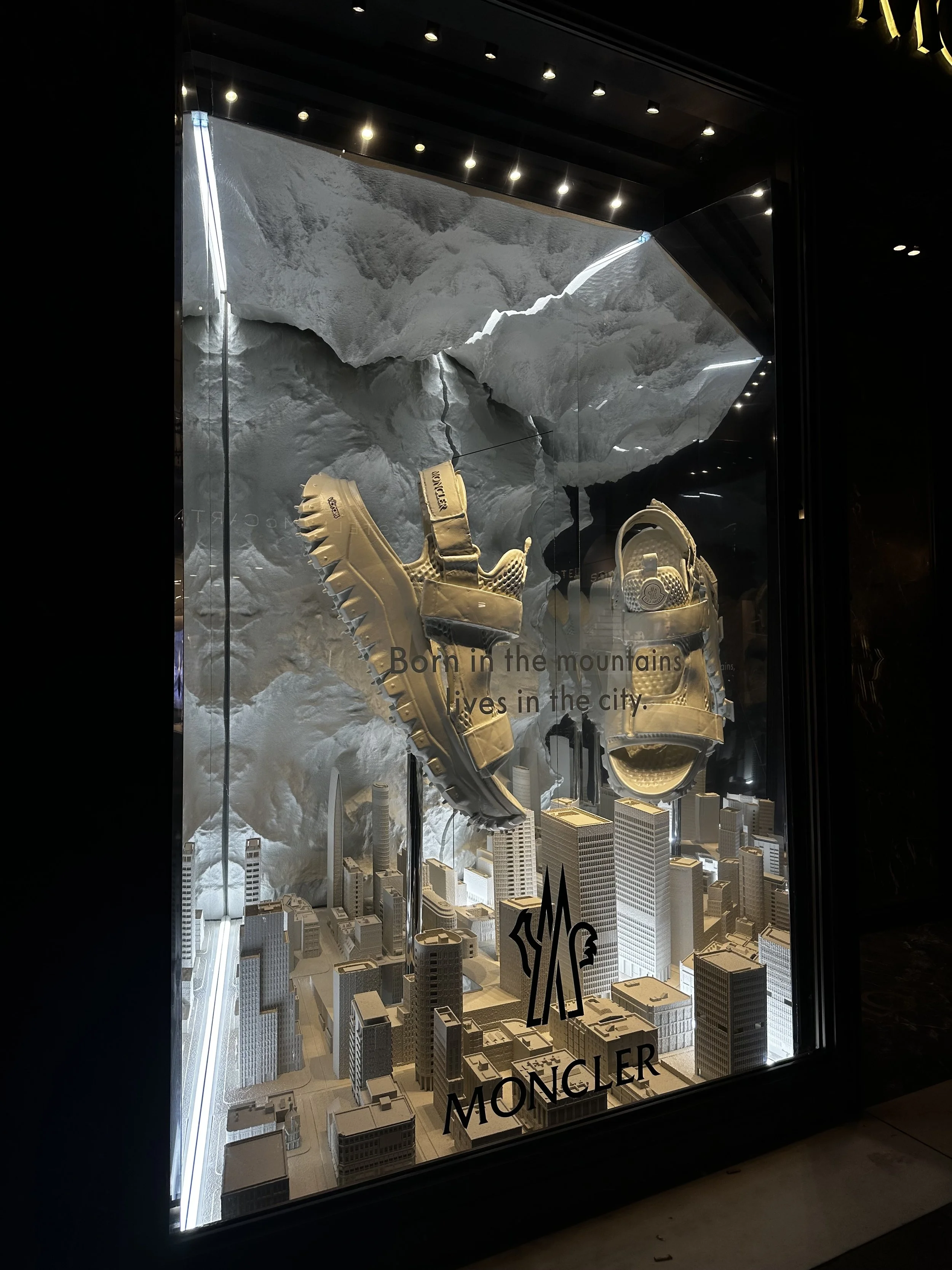 Store display featuring miniature cityscape with tall buildings and a large mountain model in the background. Text on display says 'Born in the mountains, lives in the city' and 'Moncler'.
