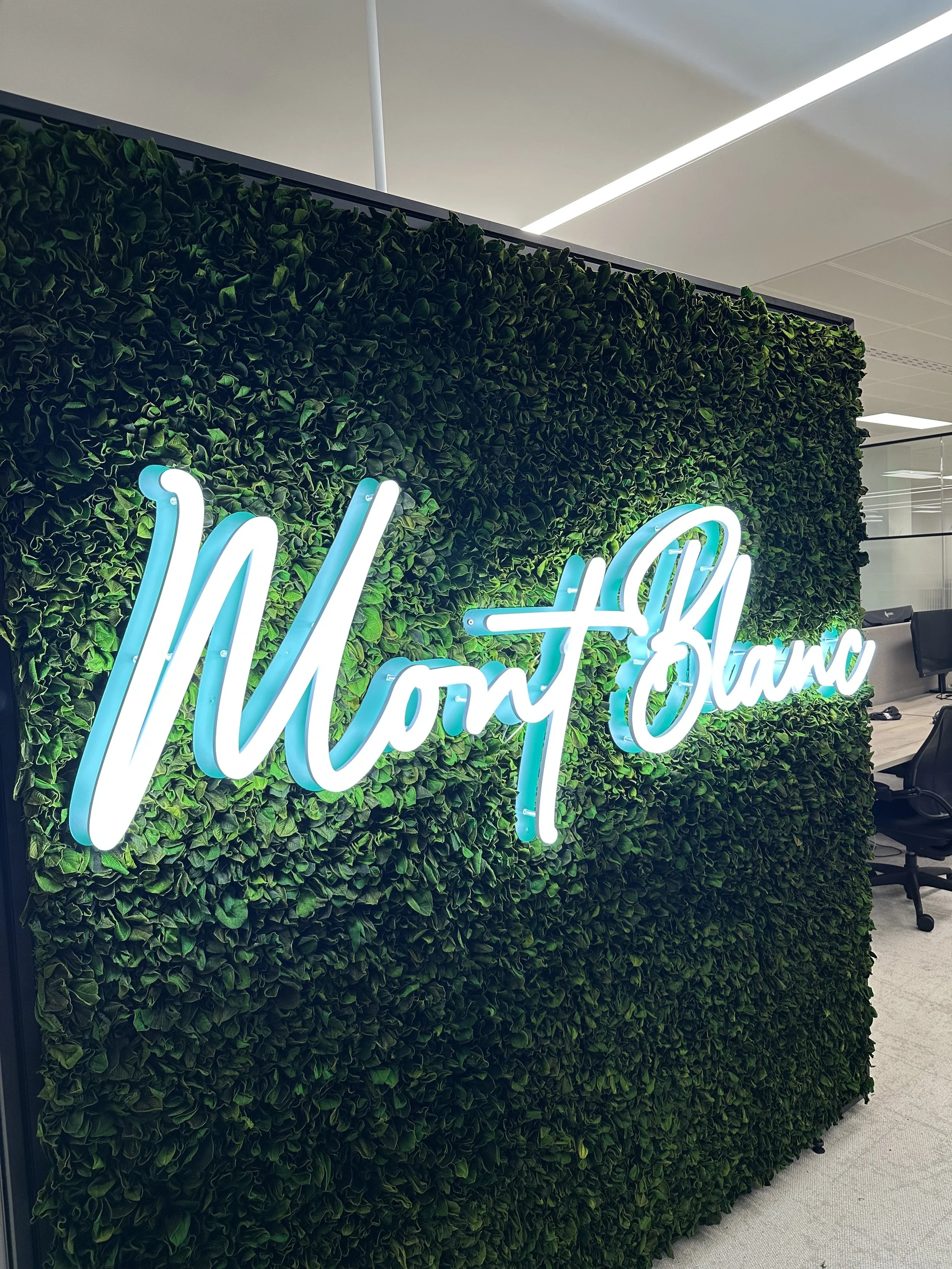 A green wall with leafy plants behind a neon sign that says "Mont Blanc" in blue cursive letters, located in an office or workspace.