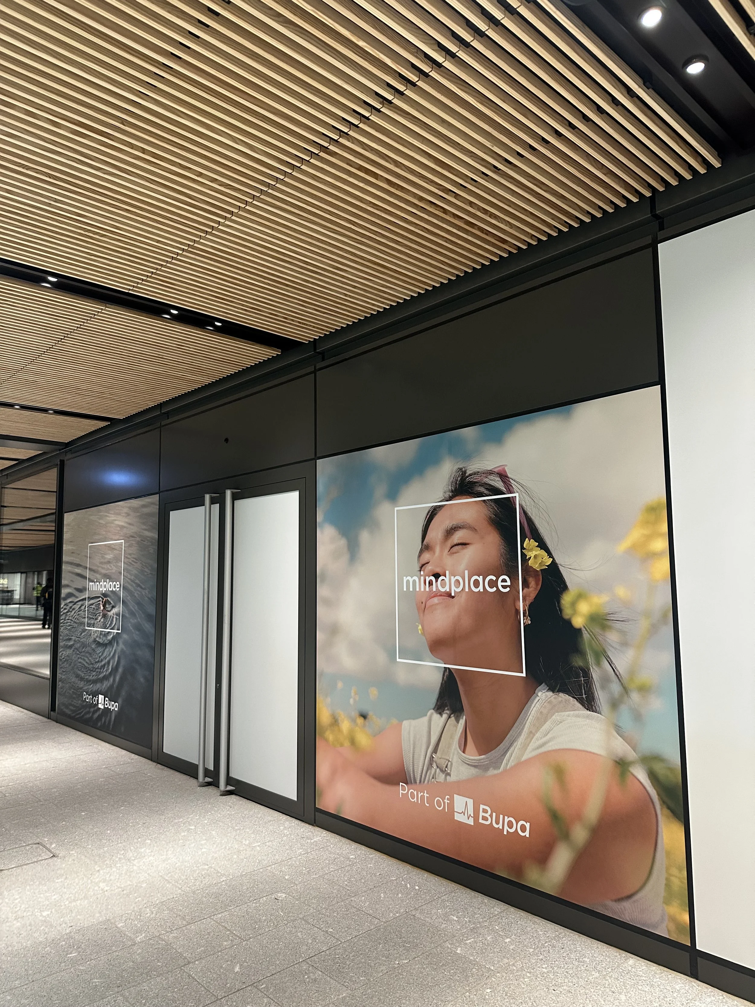window graphics showing a woman with closed eyes, smiling, with a sunny outdoor background featuring flowers and a blue sky, promoting Bupa healthcare, with the text 'mindplace' and 'Part of Bupa'.