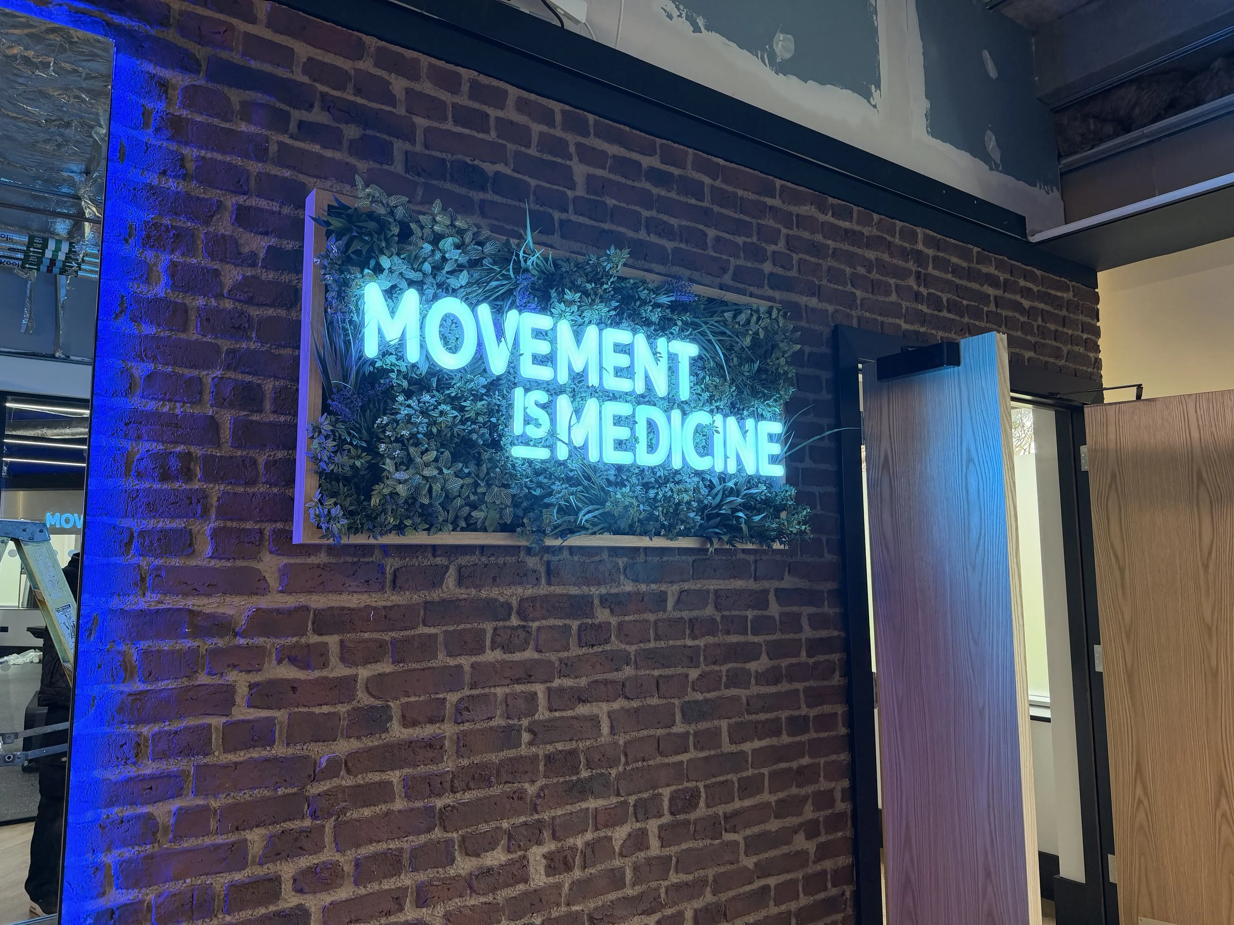 Neon blue sign reading 'Movement is Medicine' mounted on a brick wall with decorative plants around it, in an indoor space.