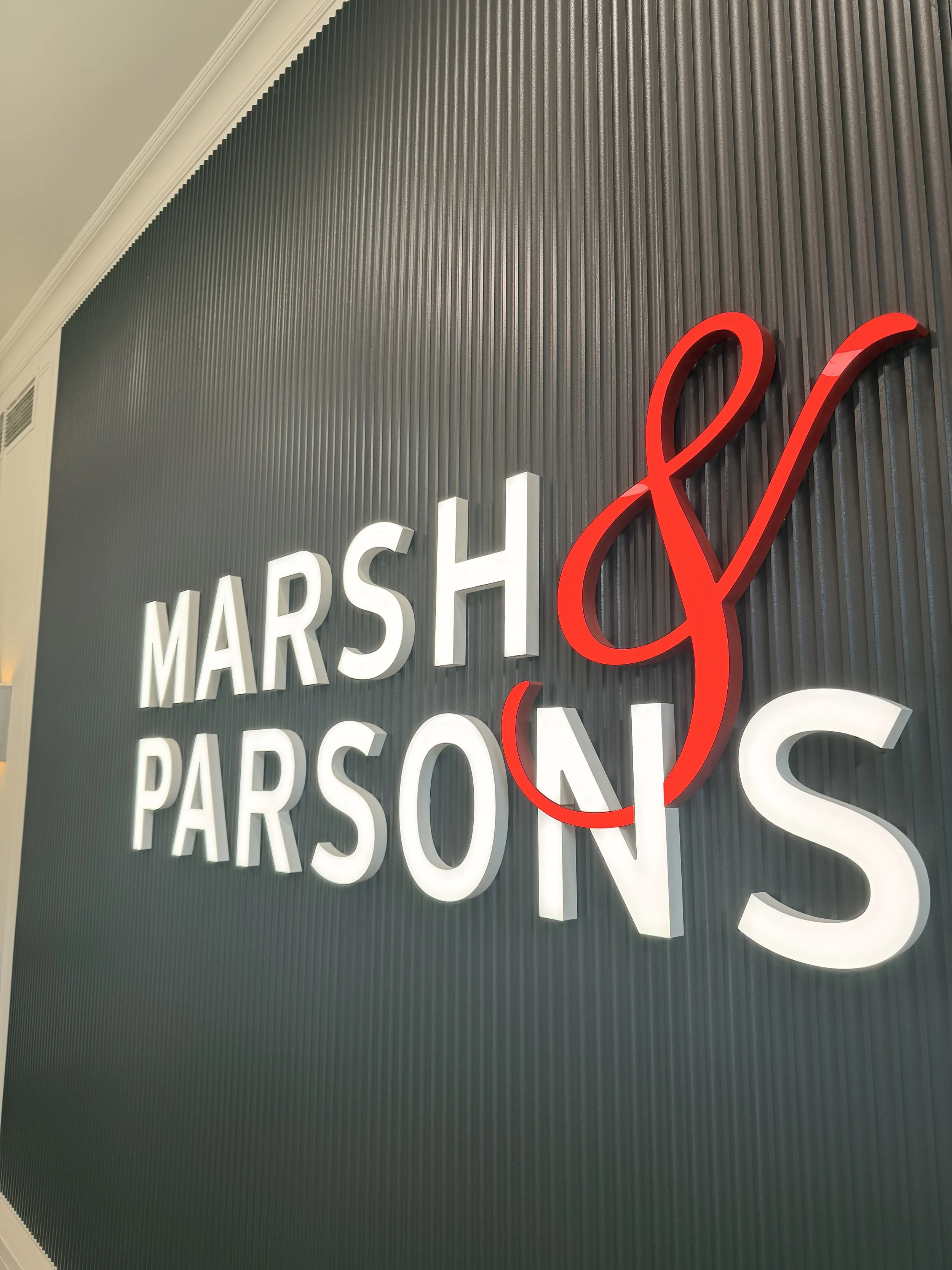Close-up of a modern wall with illuminated white and red letters spelling out 'Marsh & Parson' against a ribbed gray background.