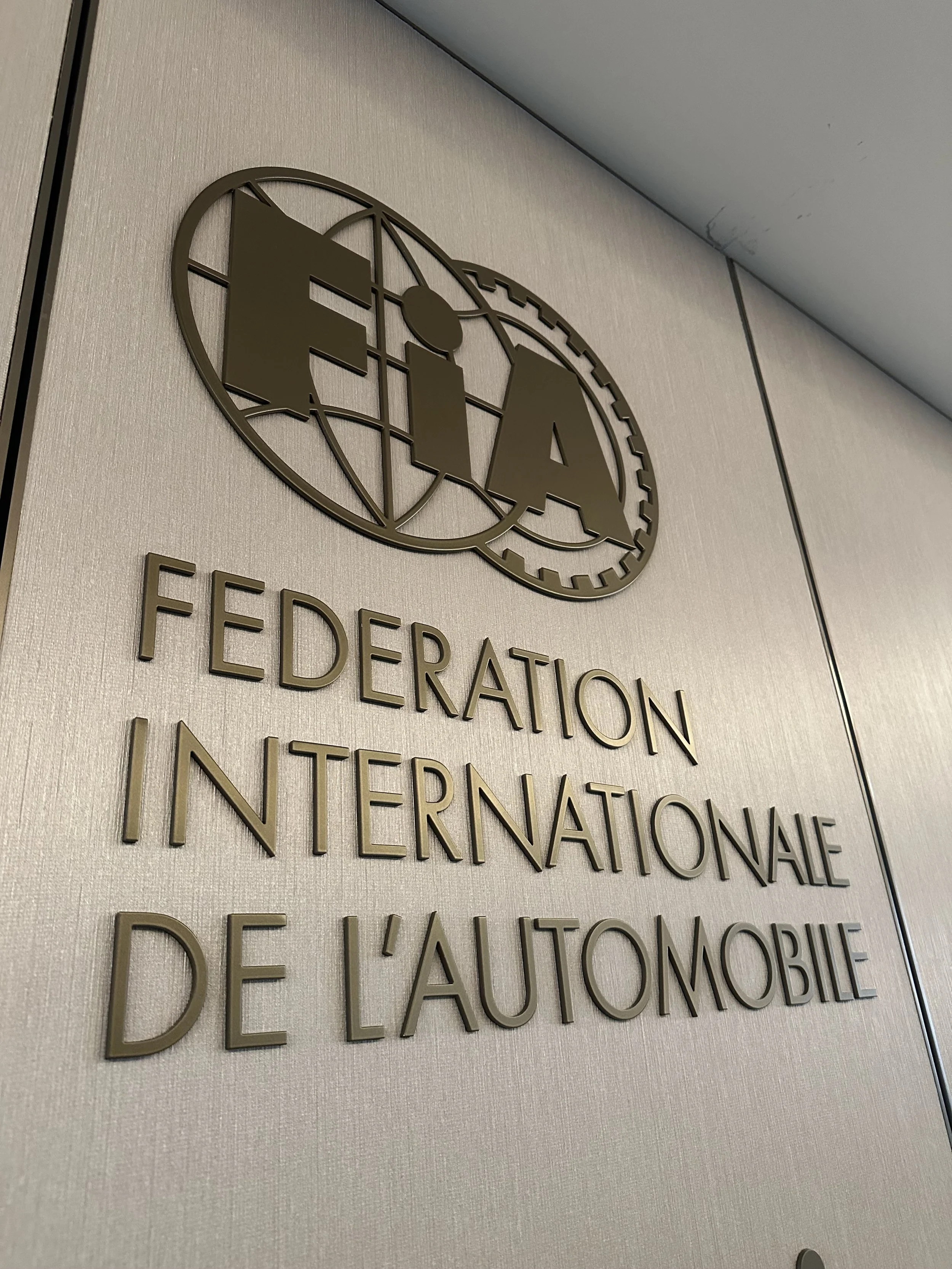 Close-up of a wall sign with the logo of the International Federation of Automation, featuring the letters 'FIA' inside an oval design with intersecting lines, and the text 'FEDERATION INTERNATIONALE DE L'AUTOMOBILE' below.