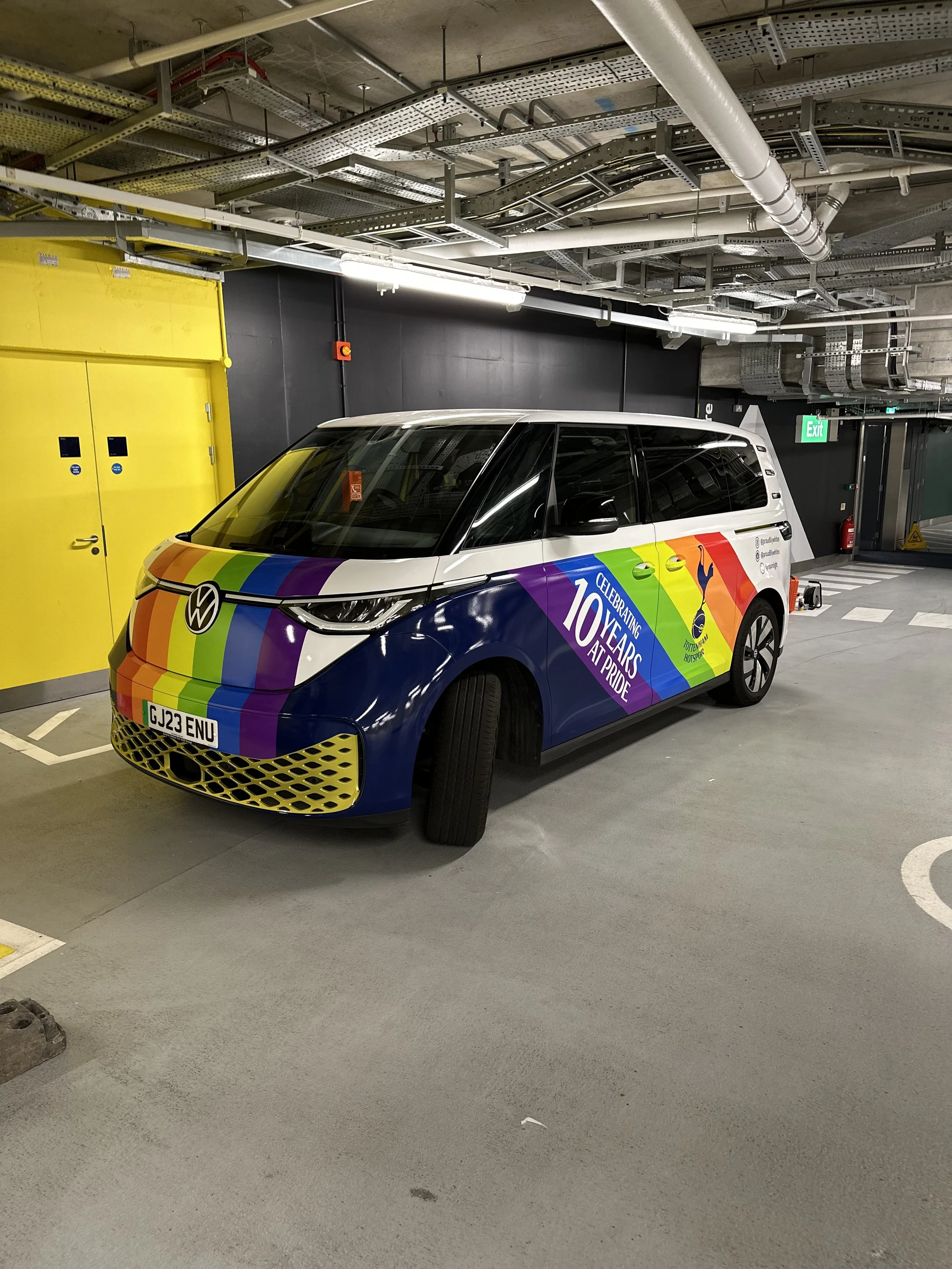 A white Volkswagen ID buzz wrapped with rainbow stripes and text for Tottenham hotspurs celebrating 10 years at Pride parked in an underground parking garage.