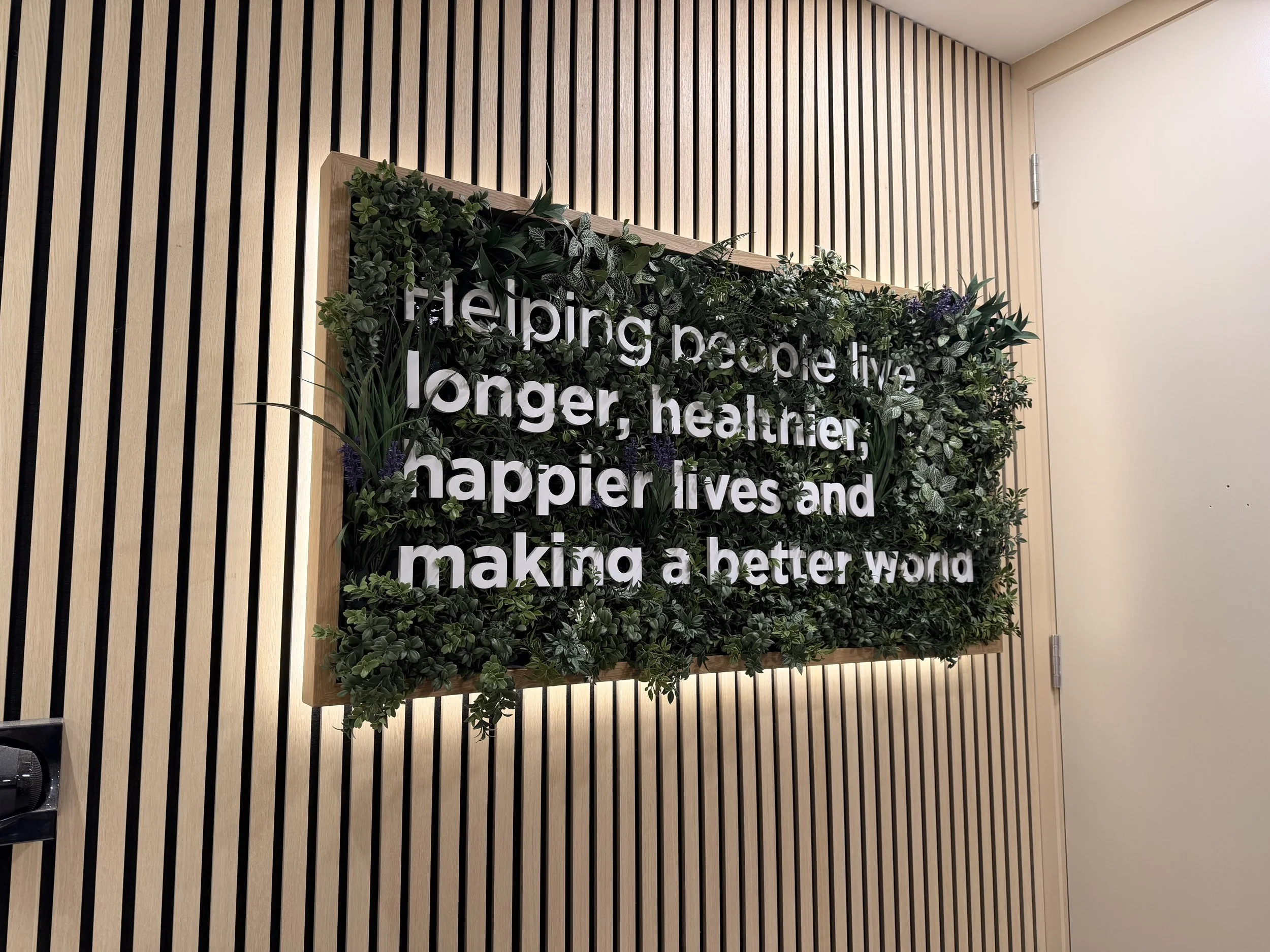 Decorative wall with vertical wooden slats and a mounted sign with greenery and white text. The greenery surrounds the sign, and part of the door and wall are visible.