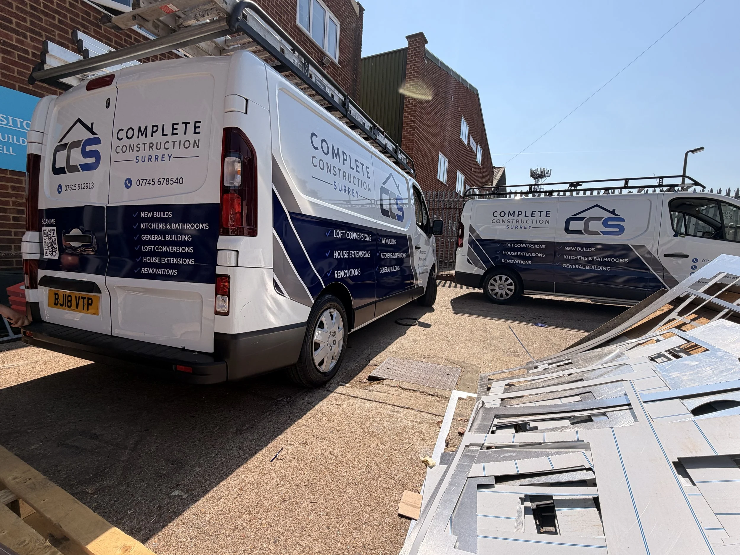 Two white vans with Complete Construction Surrey sign writing, parked outside a building under construction, with roofing materials and construction signs nearby.