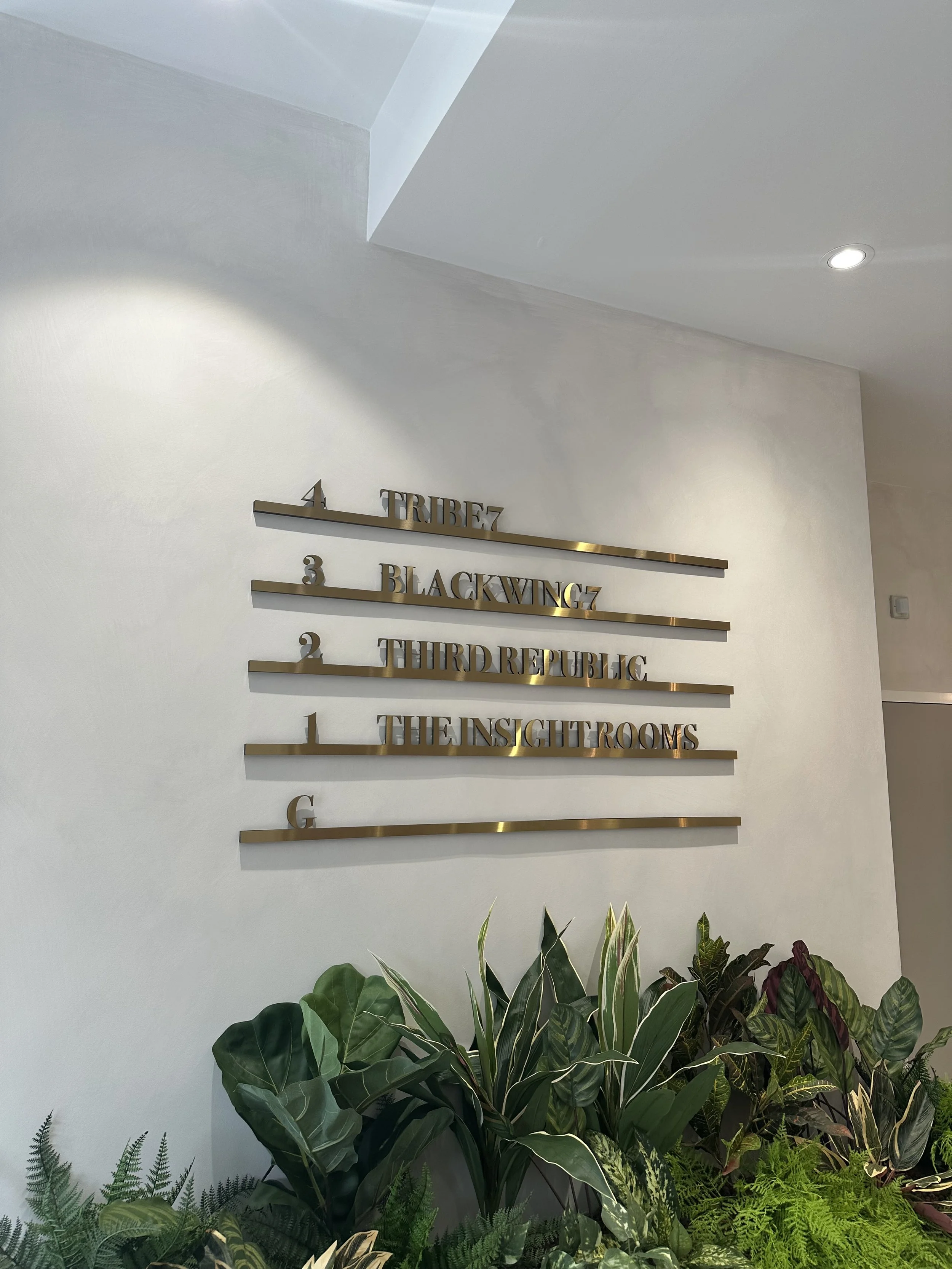 Interior wall with gold-lettered directional signs and lush green plants at the base.