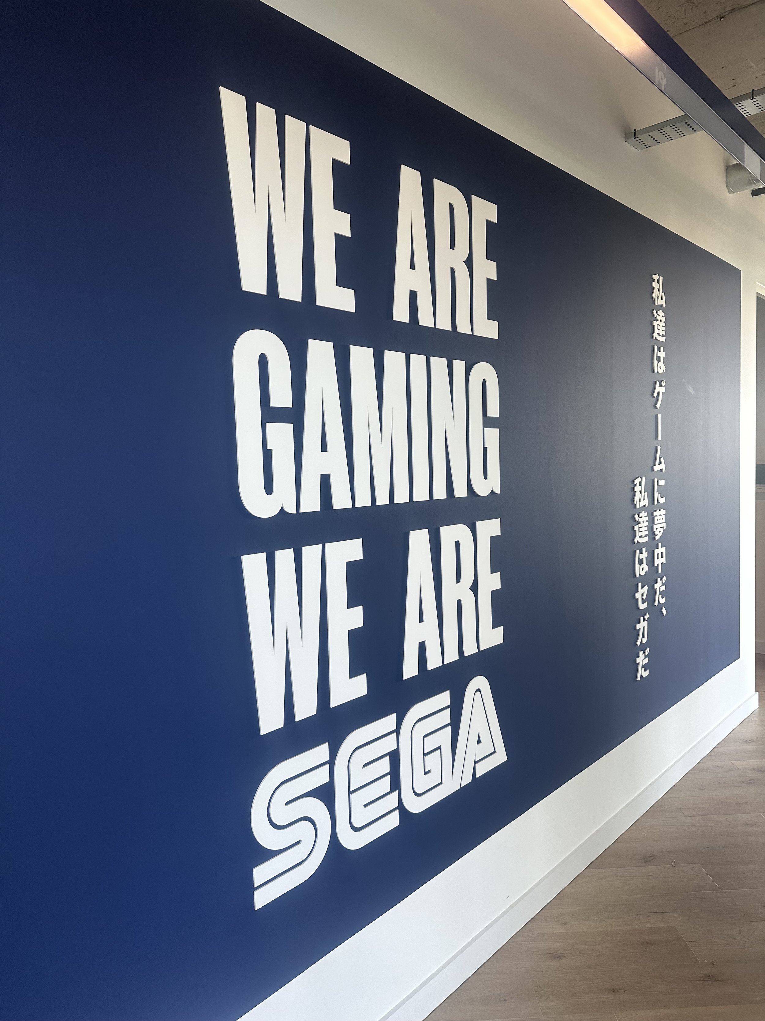 Large wall acrylic sign with bold white letters reading "WE ARE GAMING WE ARE SEGA" on a dark blue background, with Japanese characters following.