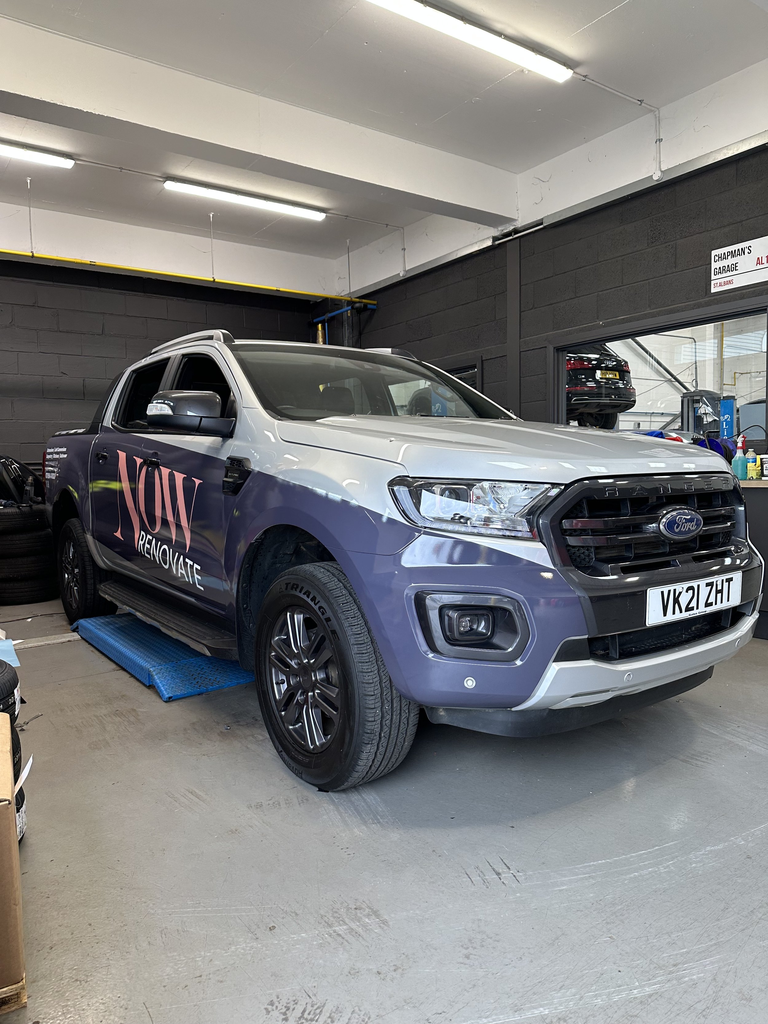 A gray Ford Ranger pickup truck parked inside an auto service garage, with a vehicle wrap for Now Renovate written on the side.