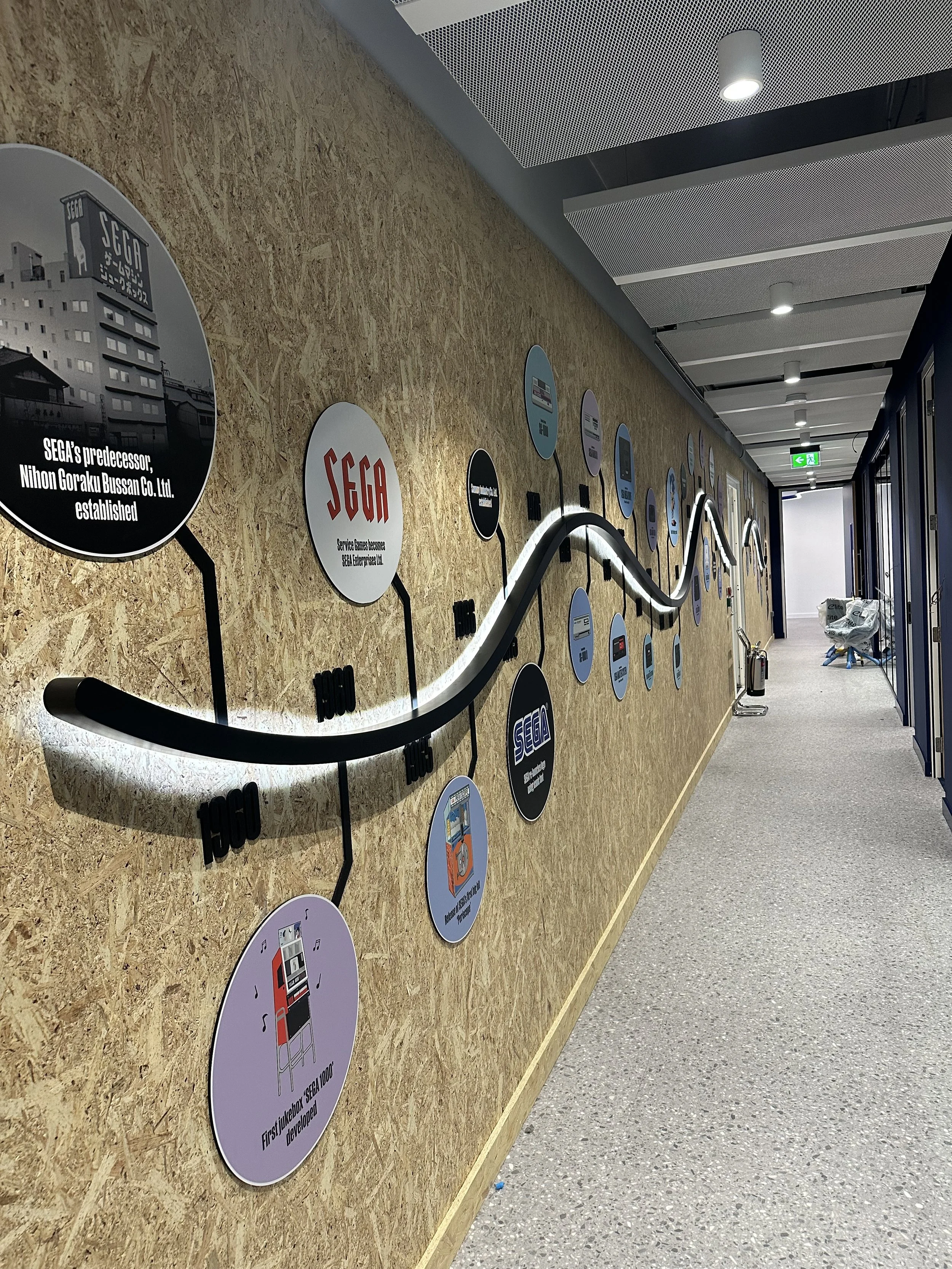 LED Wall display featuring the history of SEGA with circular graphics and a curved light strip, located in a modern corridor with ceiling lights and an emergency exit sign.